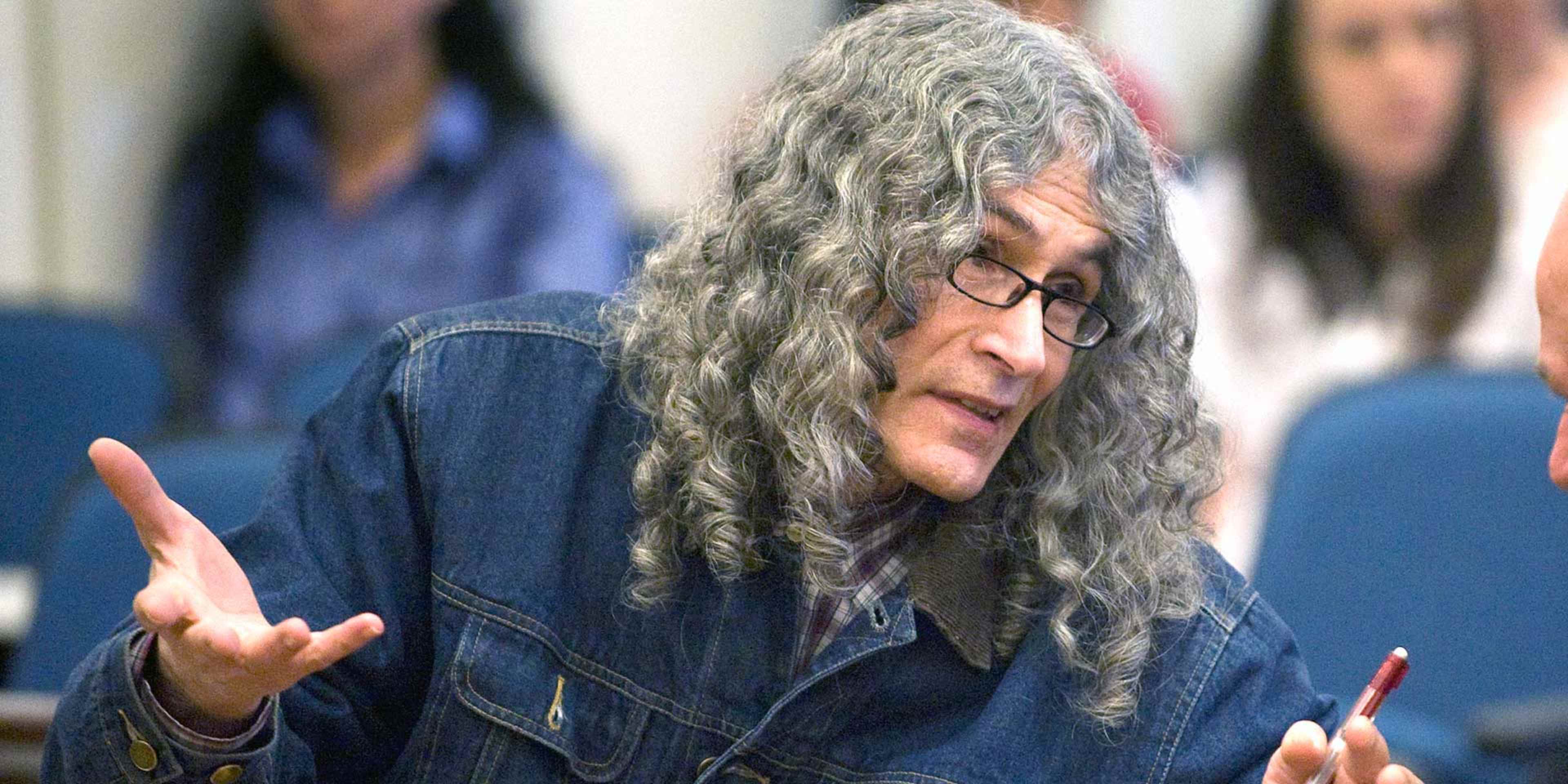 The image shows an elderly individual with long, curly gray hair and glasses, gesturing with their hands while appearing to be in conversation or discussion.