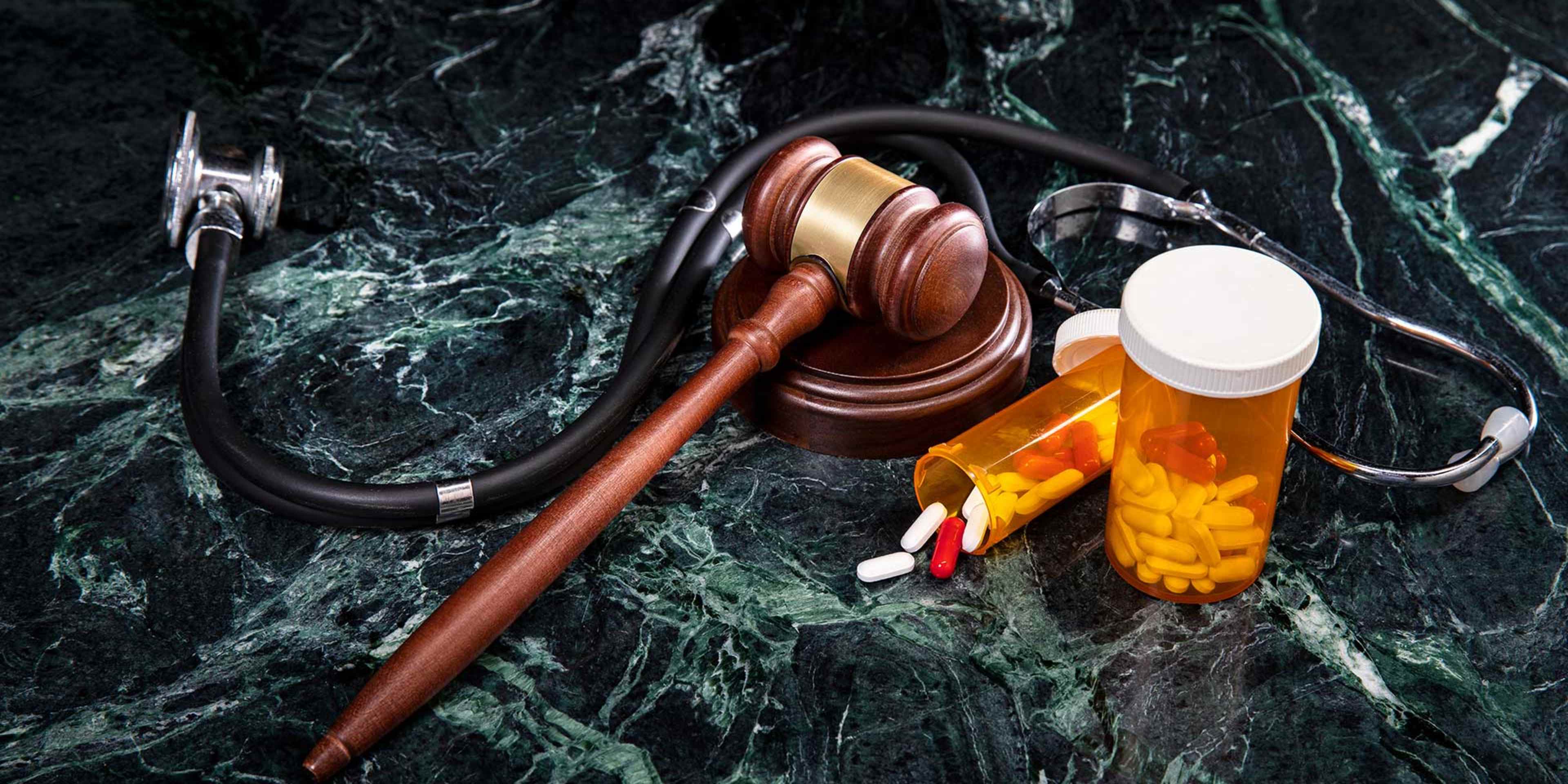 A wooden gavel, a stethoscope, and prescription medication bottles are arranged on a dark green marble background.