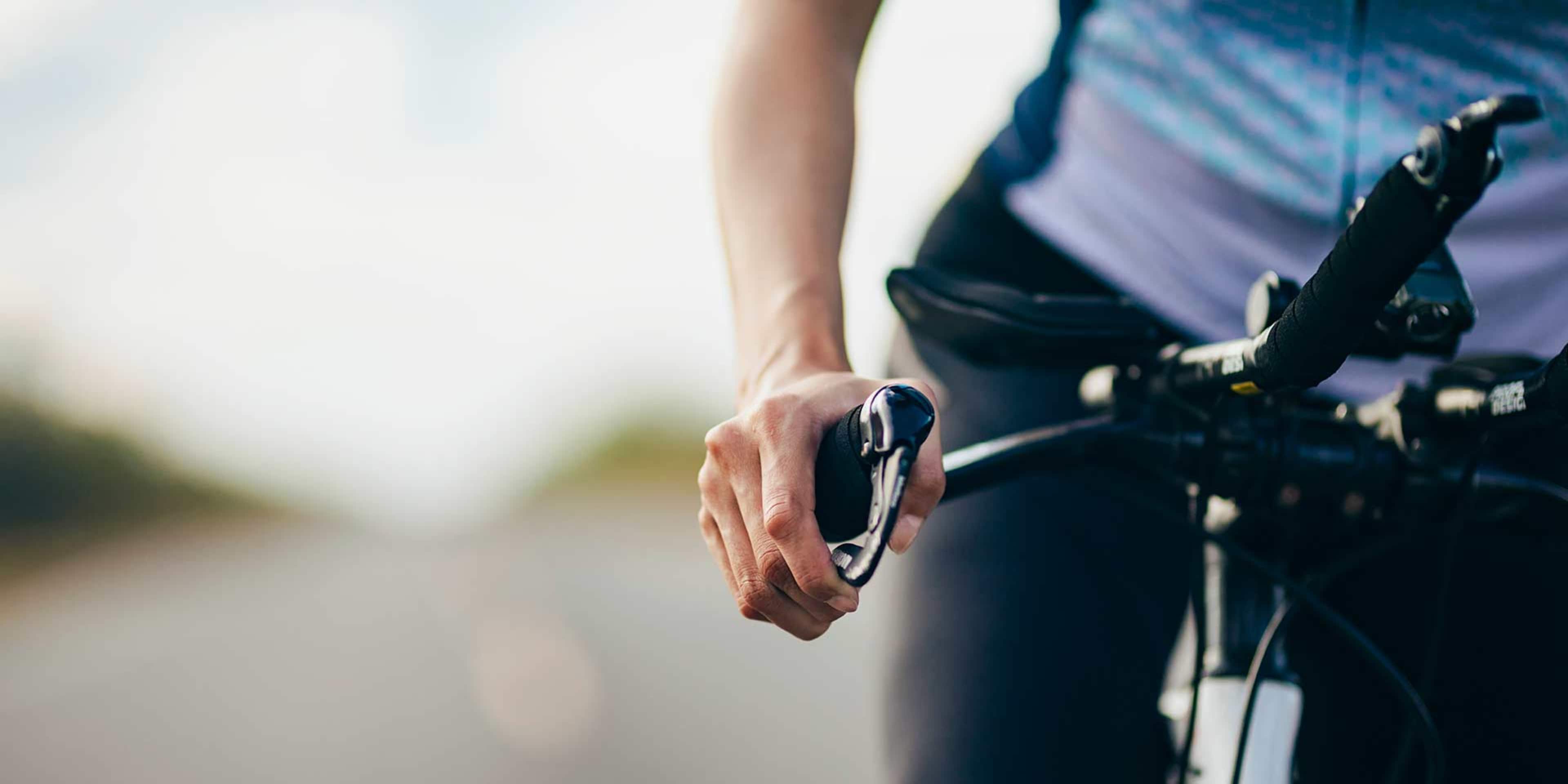 A person's hand gripping the handlebars of a bicycle, with a blurred natural landscape in the background.