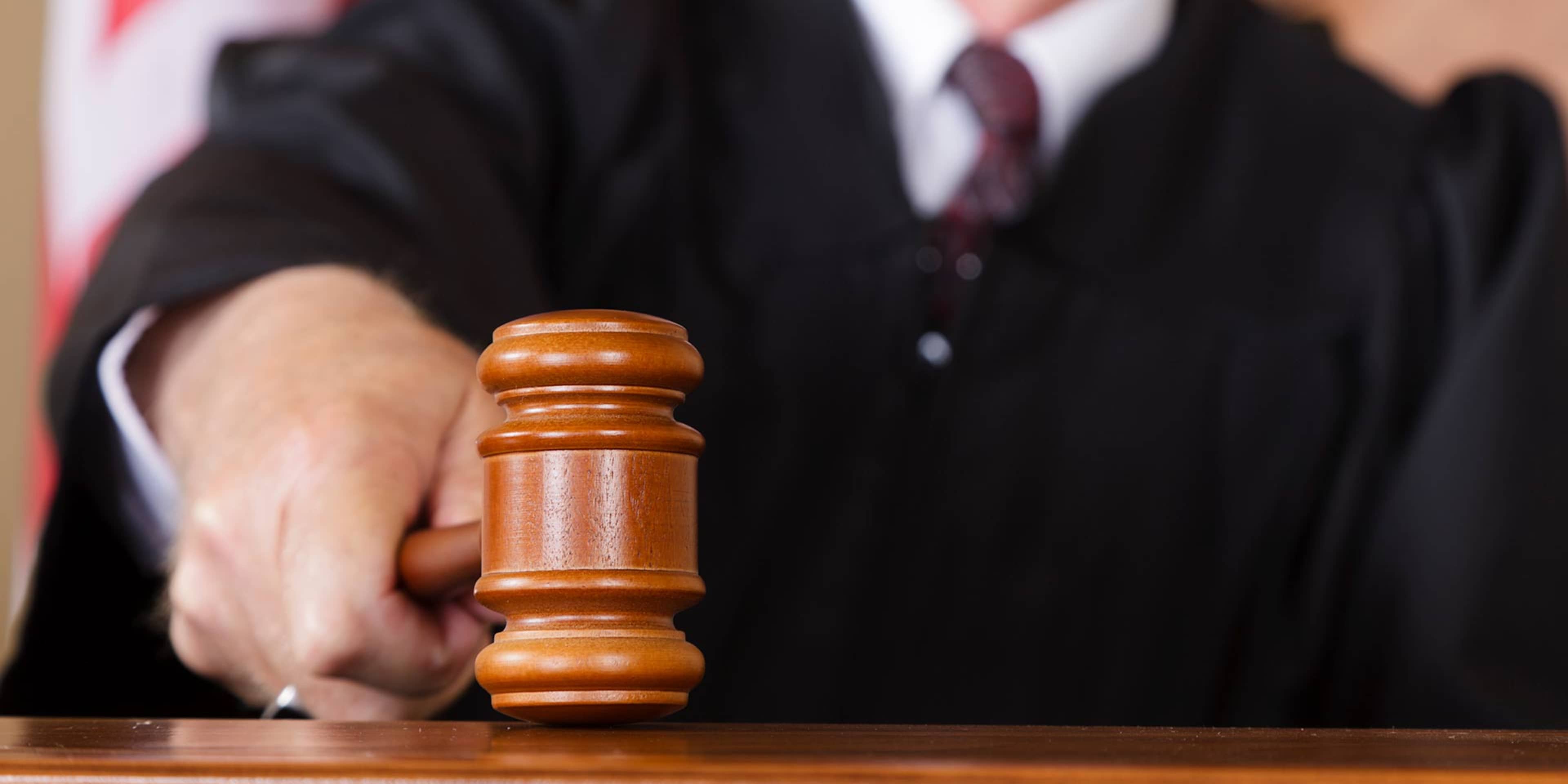 A wooden gavel is held by a person wearing a black robe, with other robed individuals visible in the background.