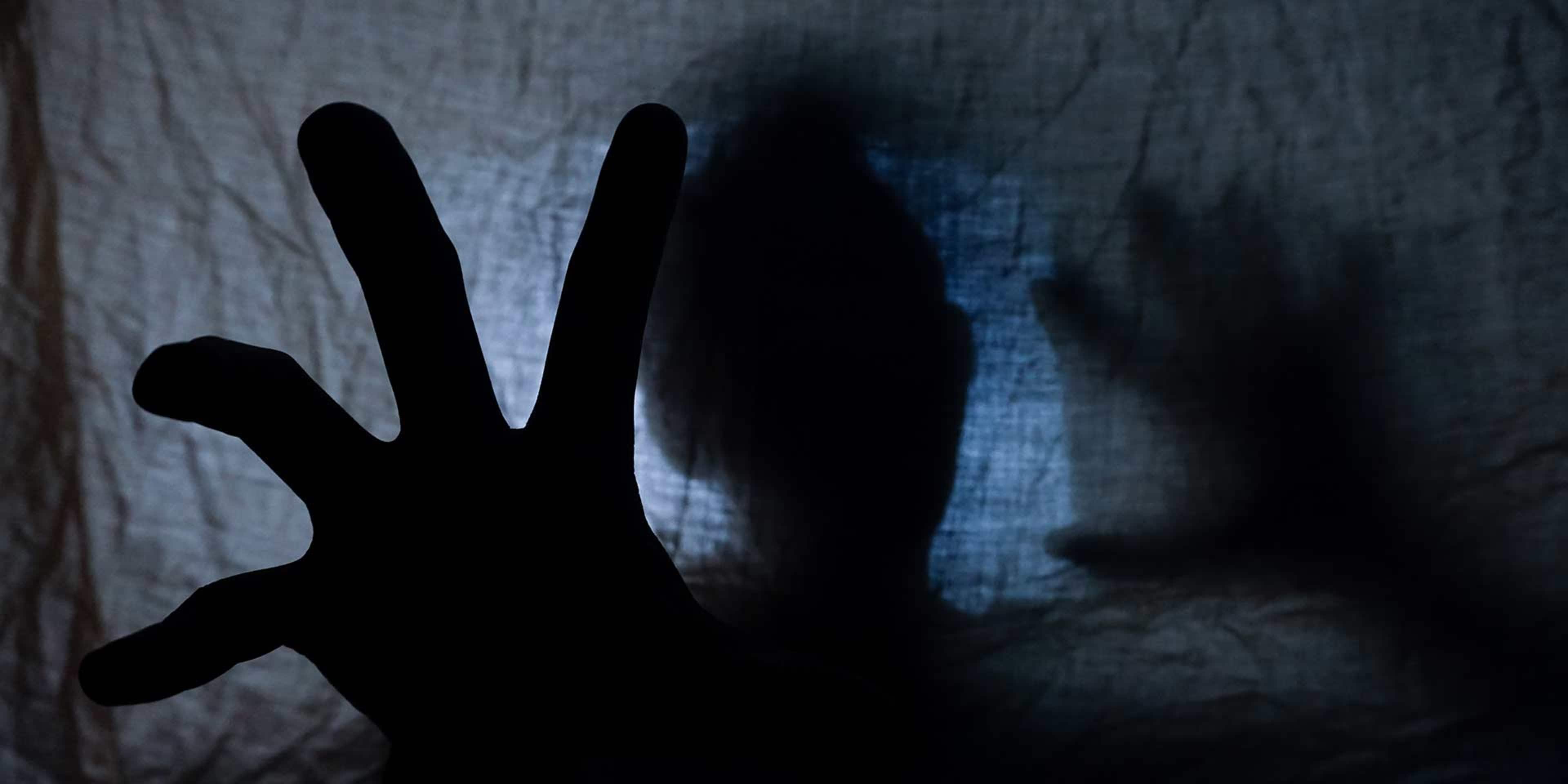 A shadowy hand gesture is visible in the foreground, set against a dark, blurred background.