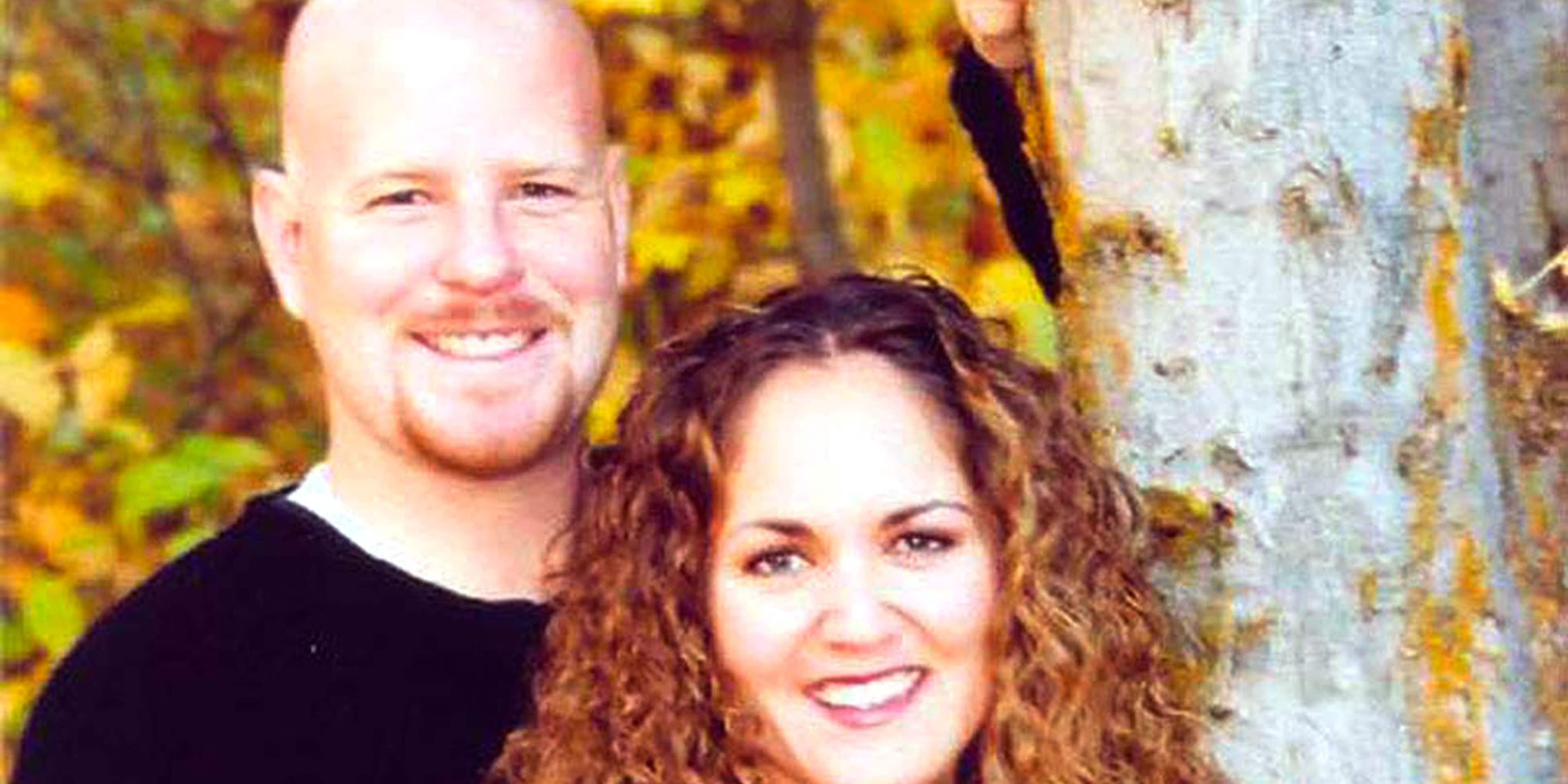 The image shows a smiling couple, a man with short gray hair and a woman with long curly red hair, standing in front of a tree with autumn foliage in the background.