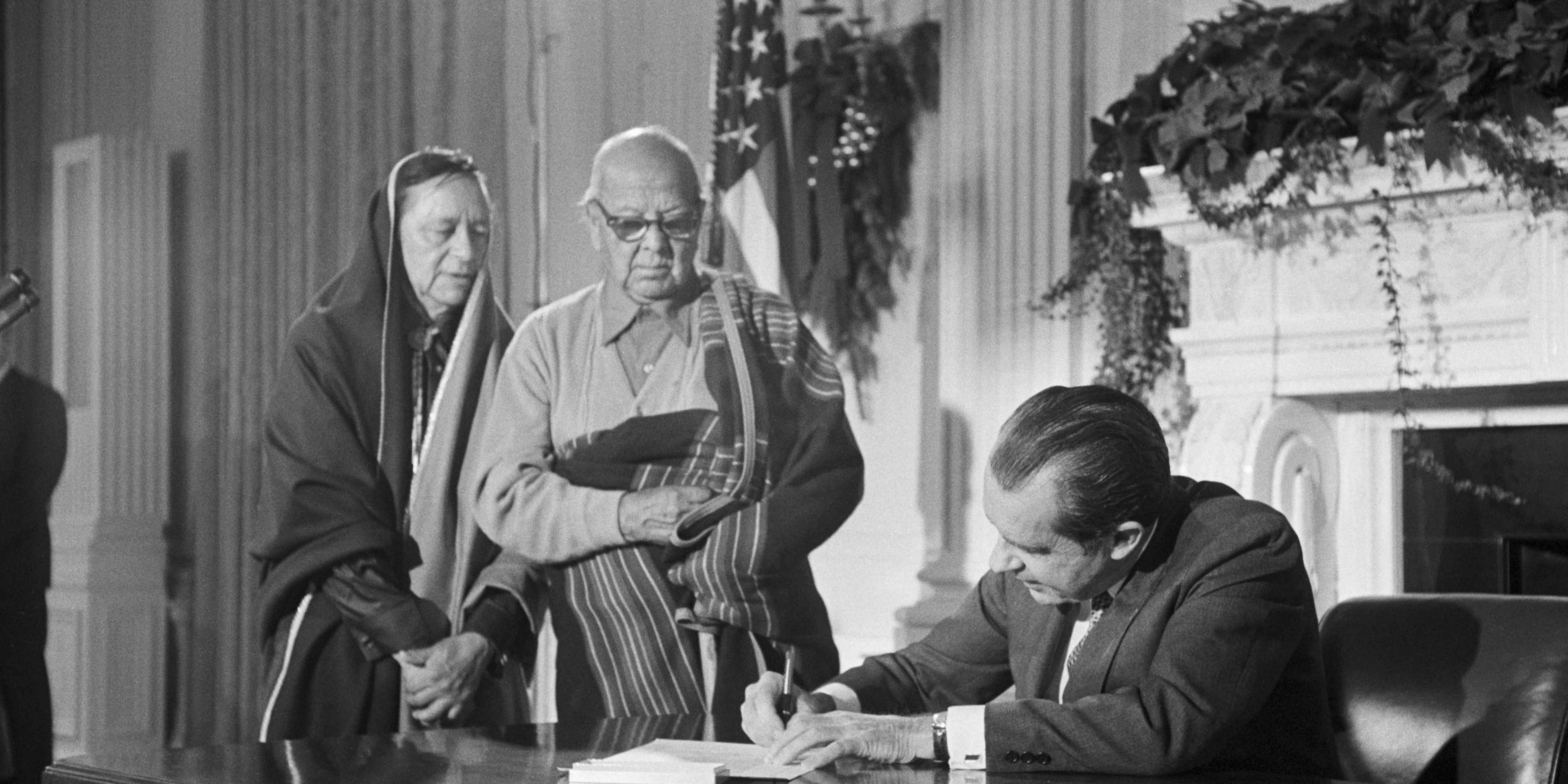 President Richard Nixon sitting at a big table signing a document while two Native American people stand to the side, looking on.