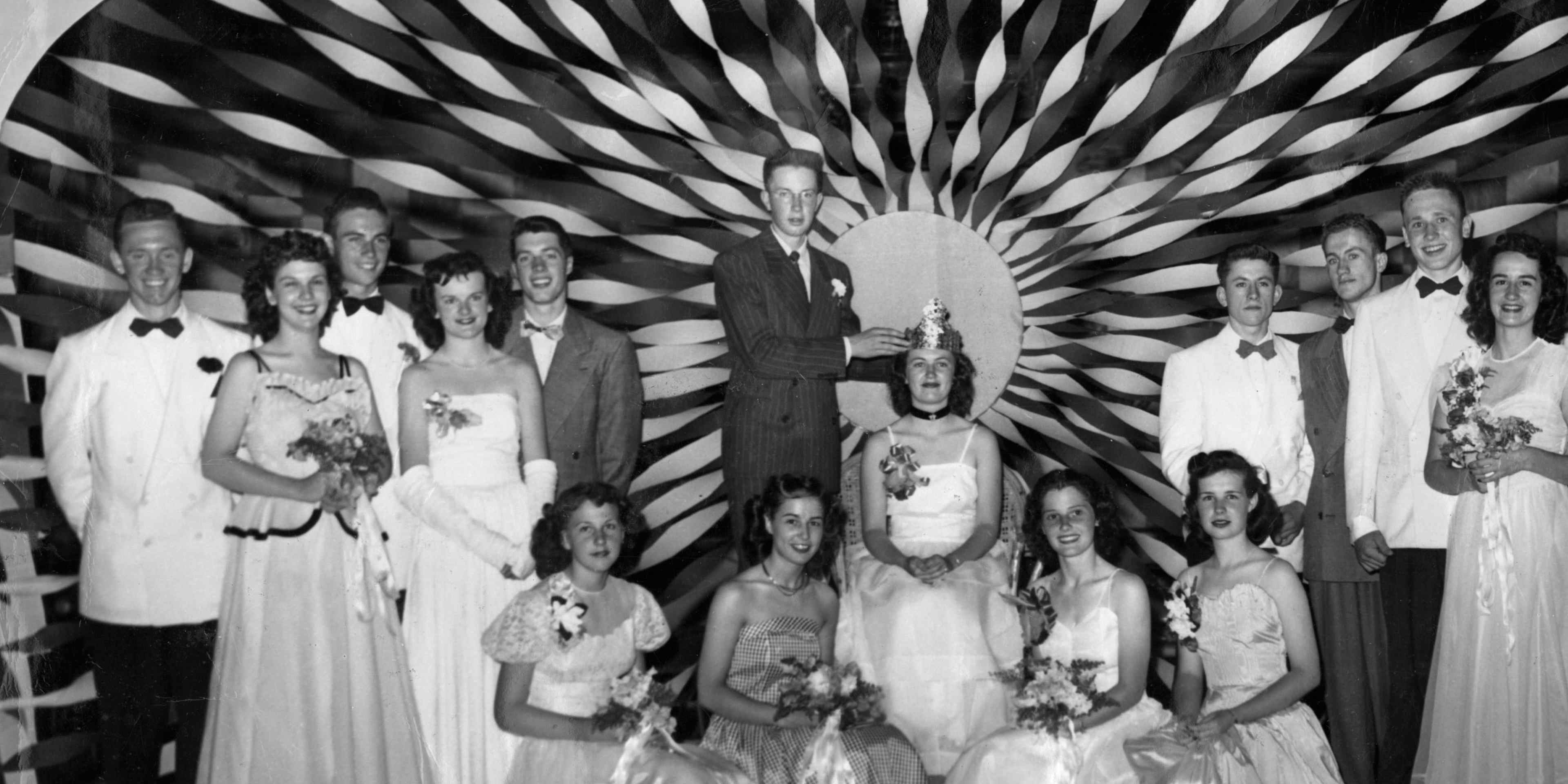 Teenagers dressed in long prom dresses and suits crowning the prom queen, with streamers radiating behind them like the rays of the sun