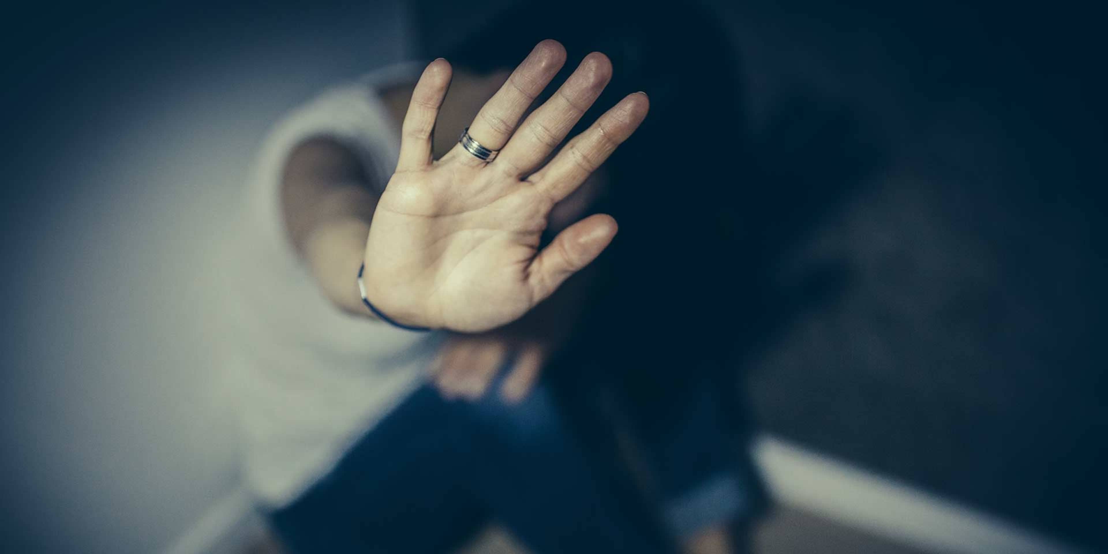 A hand is raised in a gesture of defense or protection against a dark, blurred background.