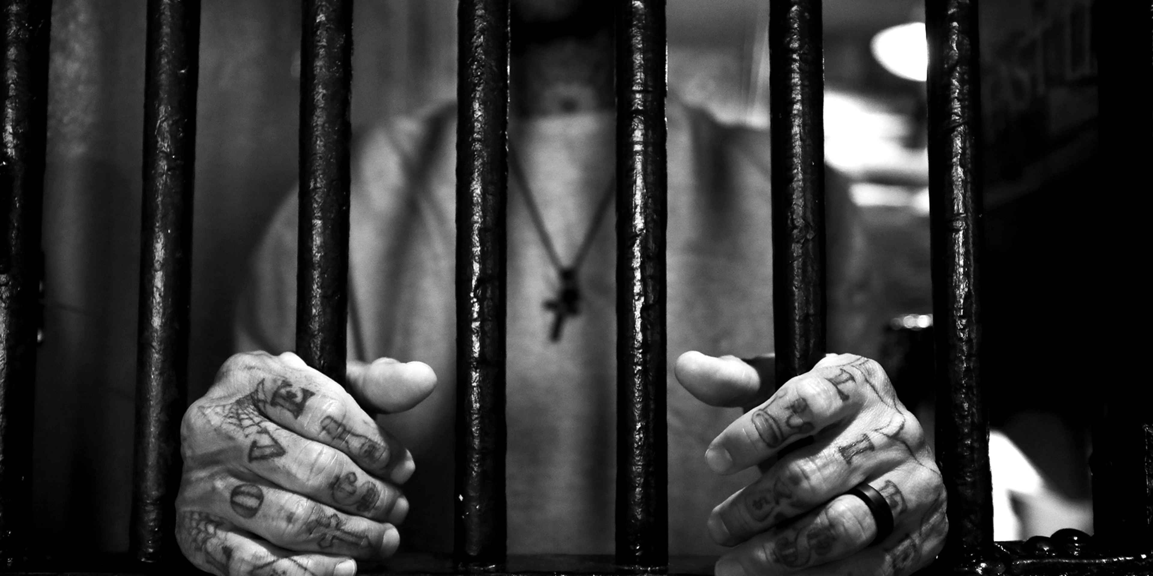 Hands grasping the bars of a prison cell, with a blurred figure visible in the background.
