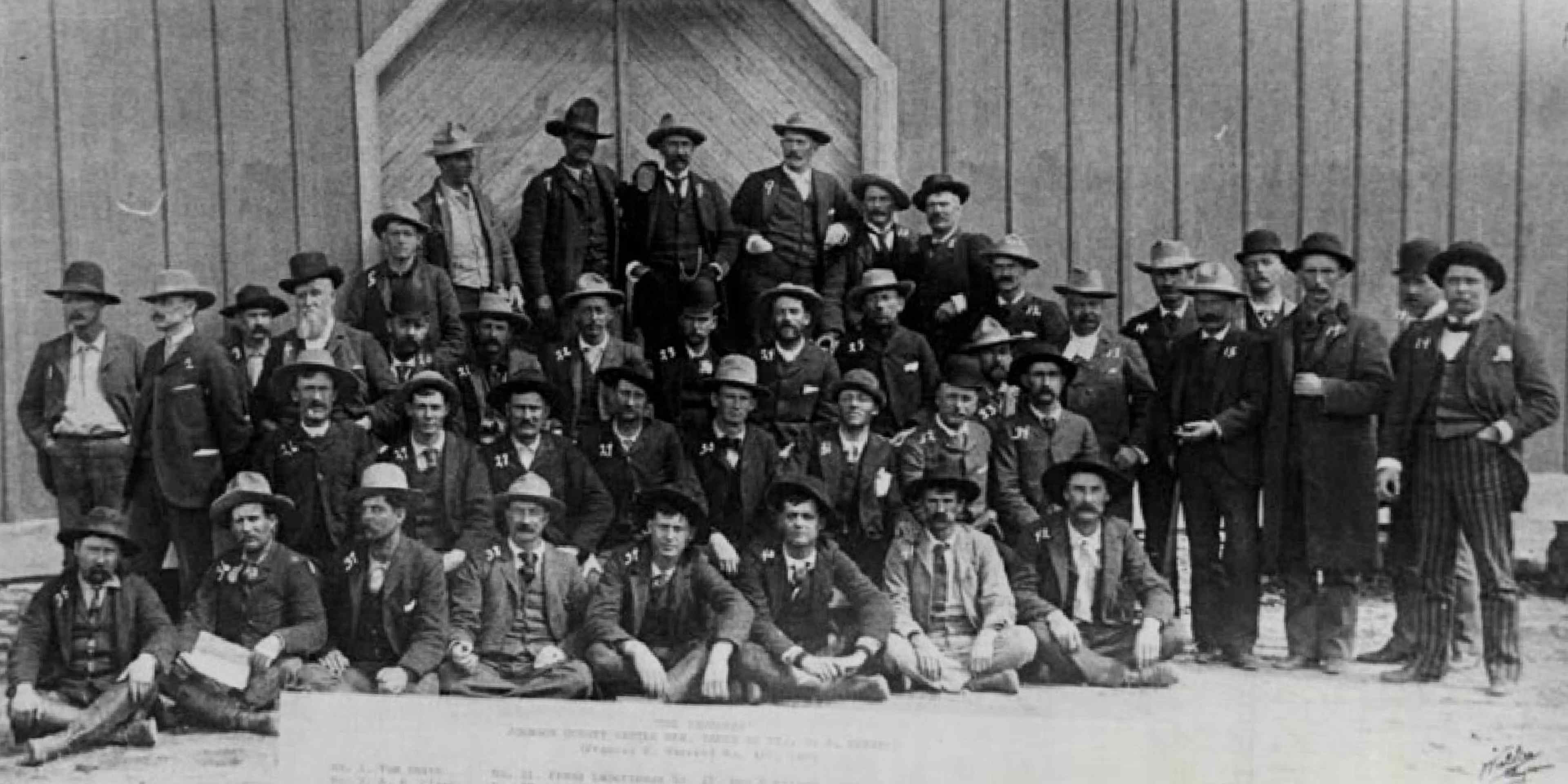 About 40 men in suits and hats posed in front of a barn. After this group of hitmen hired by Wyoming cattle barons to murder settlers were rescued from a siege by the cavalry, they were lined up for this photo. Due to influence from their wealthy backers, the gunmen were all eventually released.