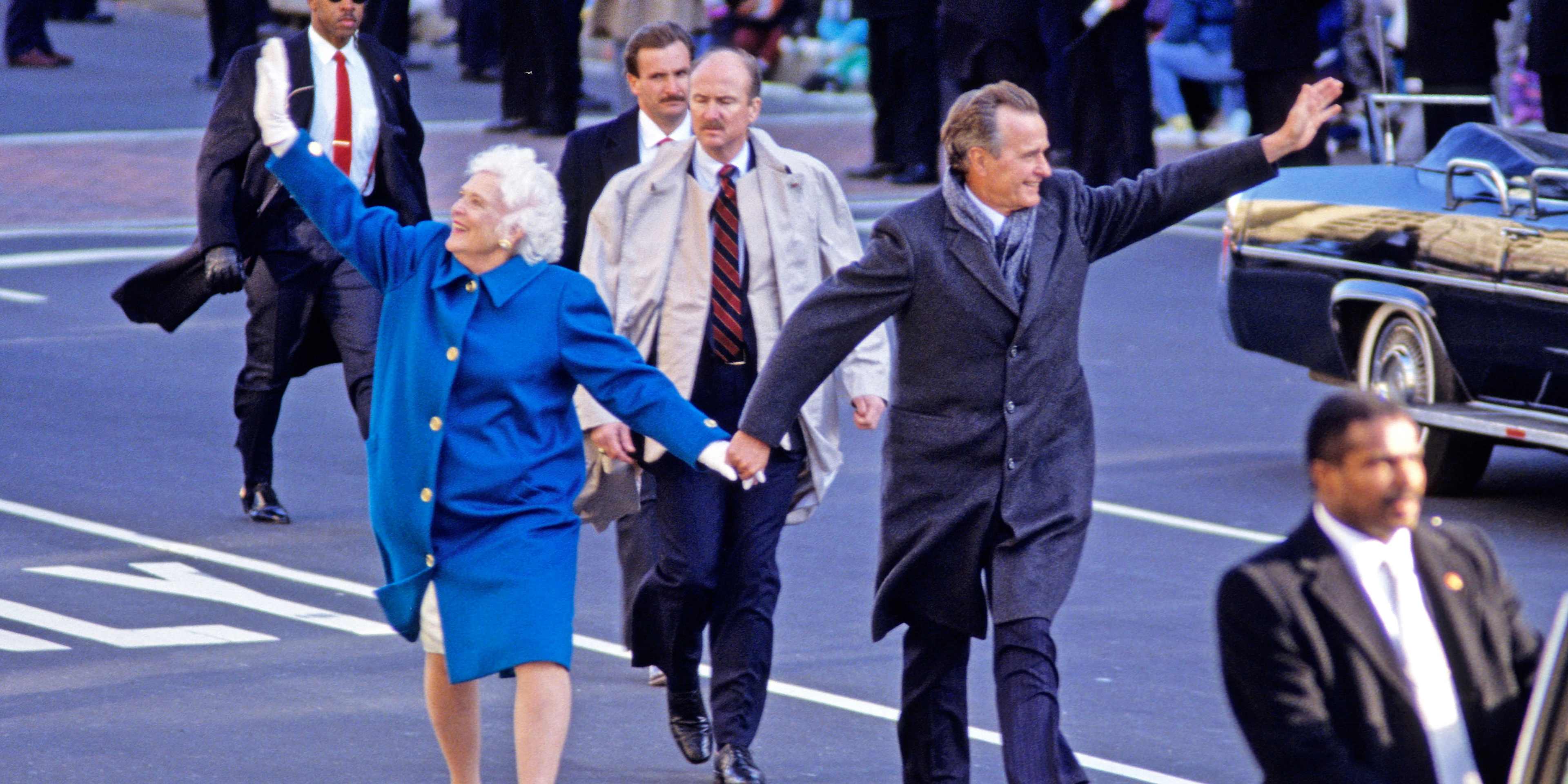 First Lady Barbara Bush (wearing a bright blue coat) and US President George HW Bush (in a gray overcoat and scarf) hold hands, smile and wave as they lead the Inaugural Parade along Pennsylvania Avenue, Washington DC, January 20, 1989. In front of and behind them in the middle of the street are various Secret Service agents.