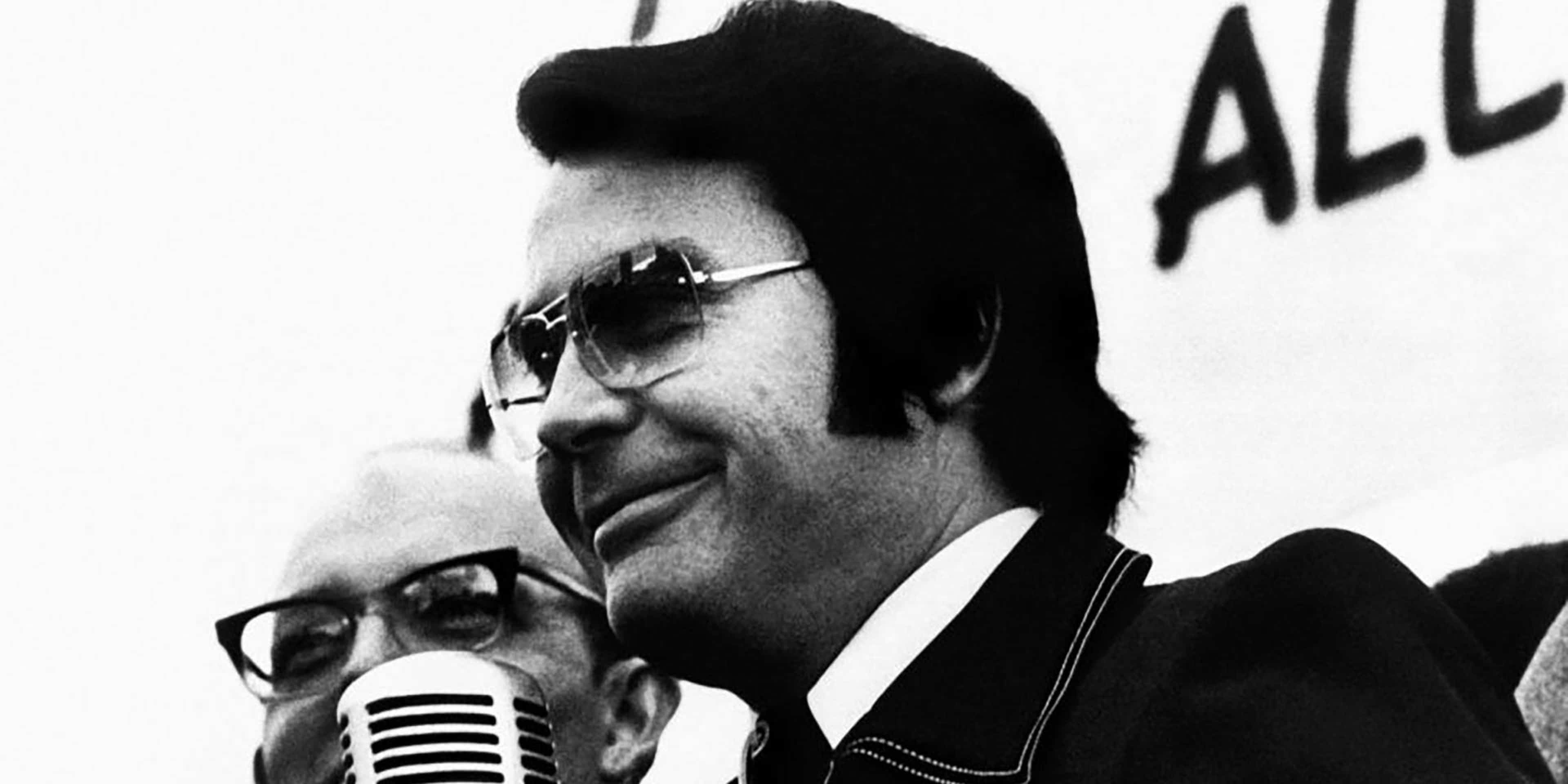 A black and white photograph captures a man wearing sunglasses and holding a microphone, with the word "ALL" prominently displayed in the background.