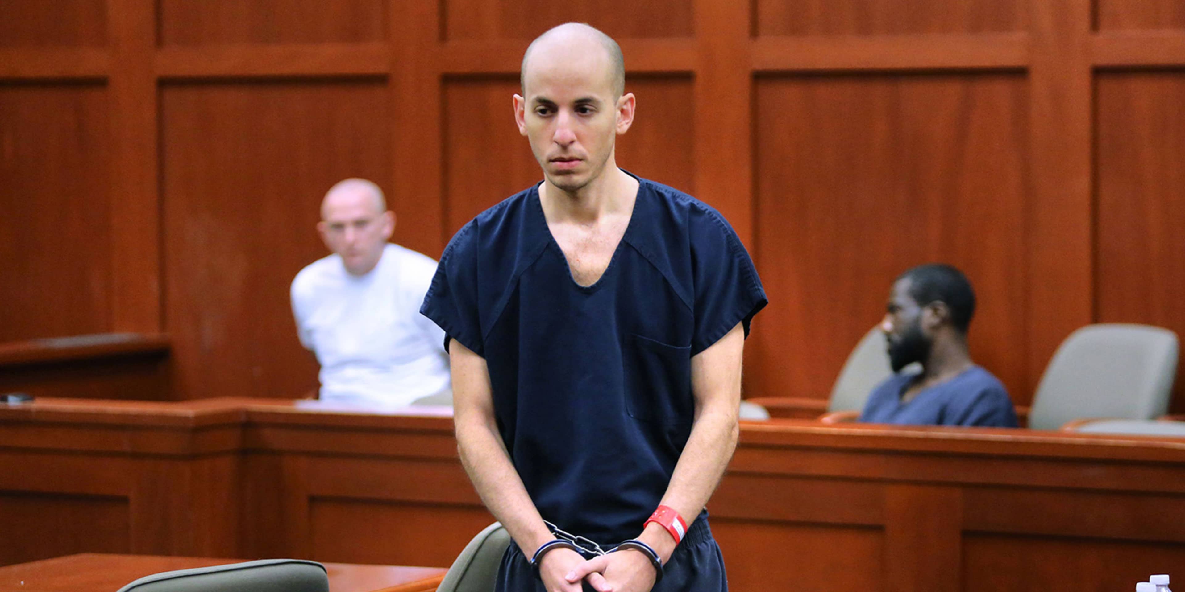 The image shows a man in a navy blue shirt standing in a courtroom, with two other individuals visible in the background.