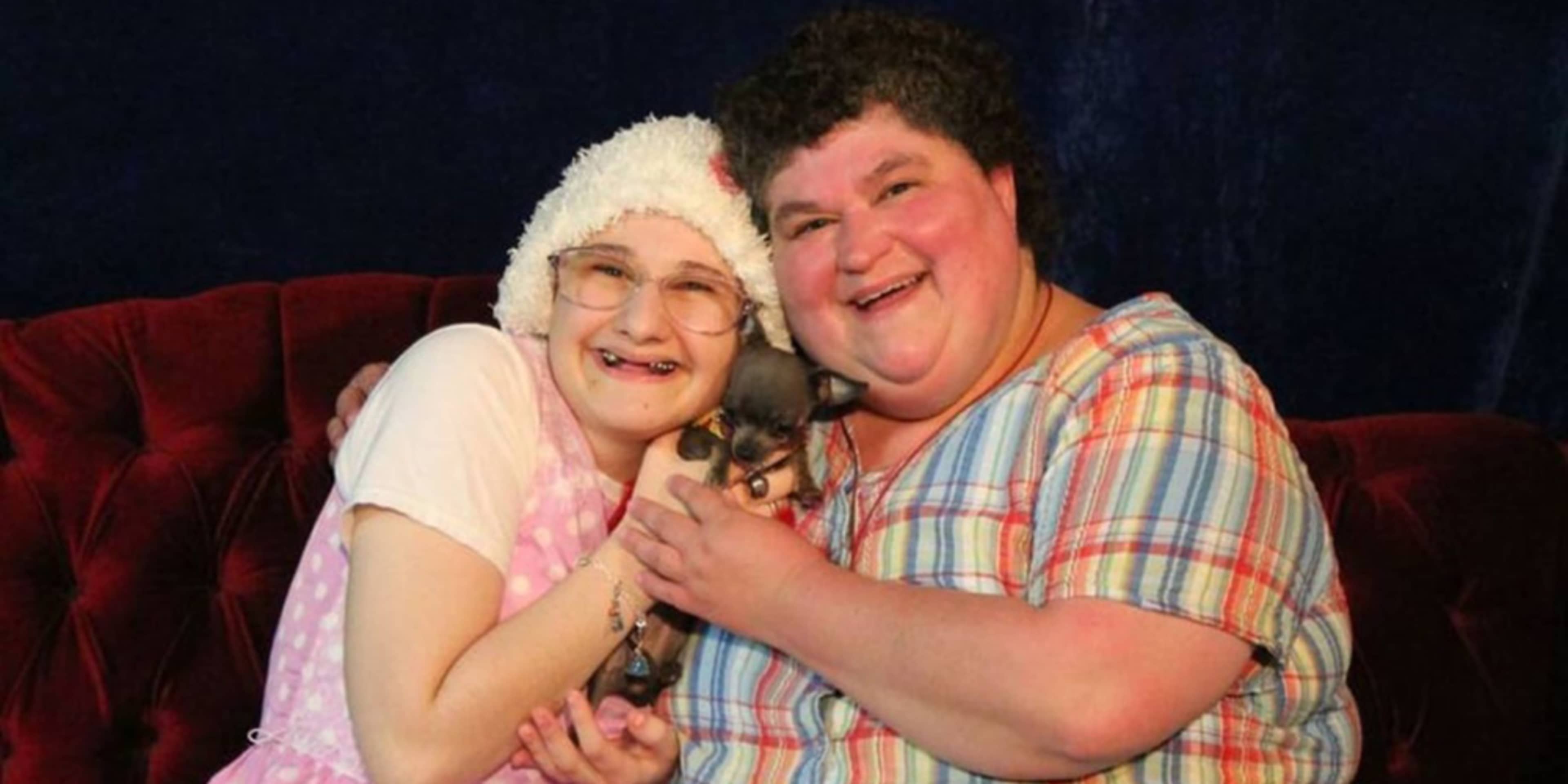 Two elderly women, one wearing a white hat and the other a plaid shirt, are embracing and smiling warmly on a red couch against a dark background.
