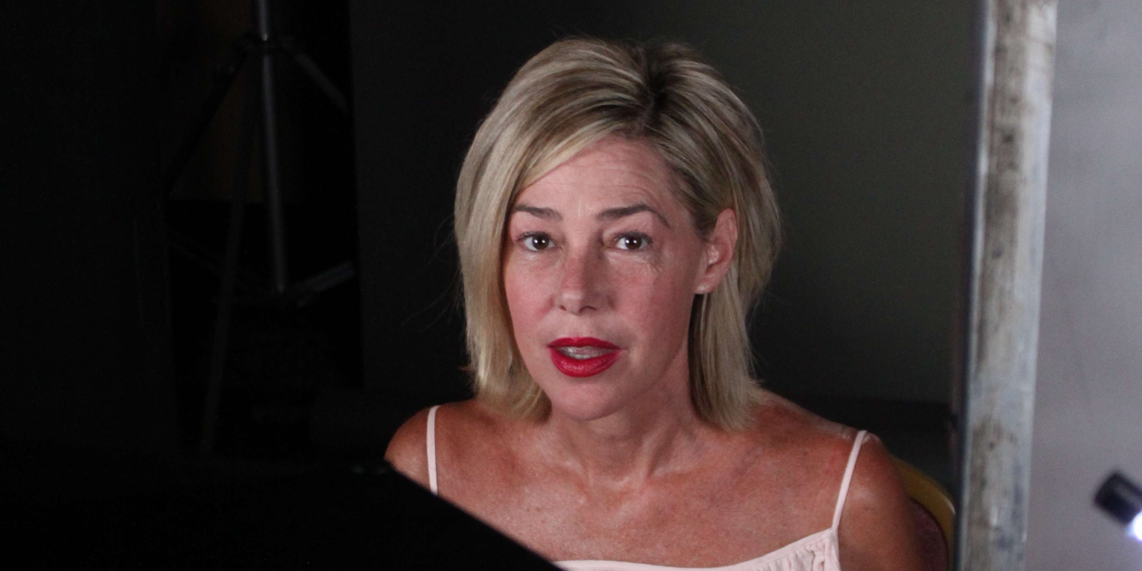 A woman with blonde hair and red lipstick appears in the foreground, with a dark background behind her.