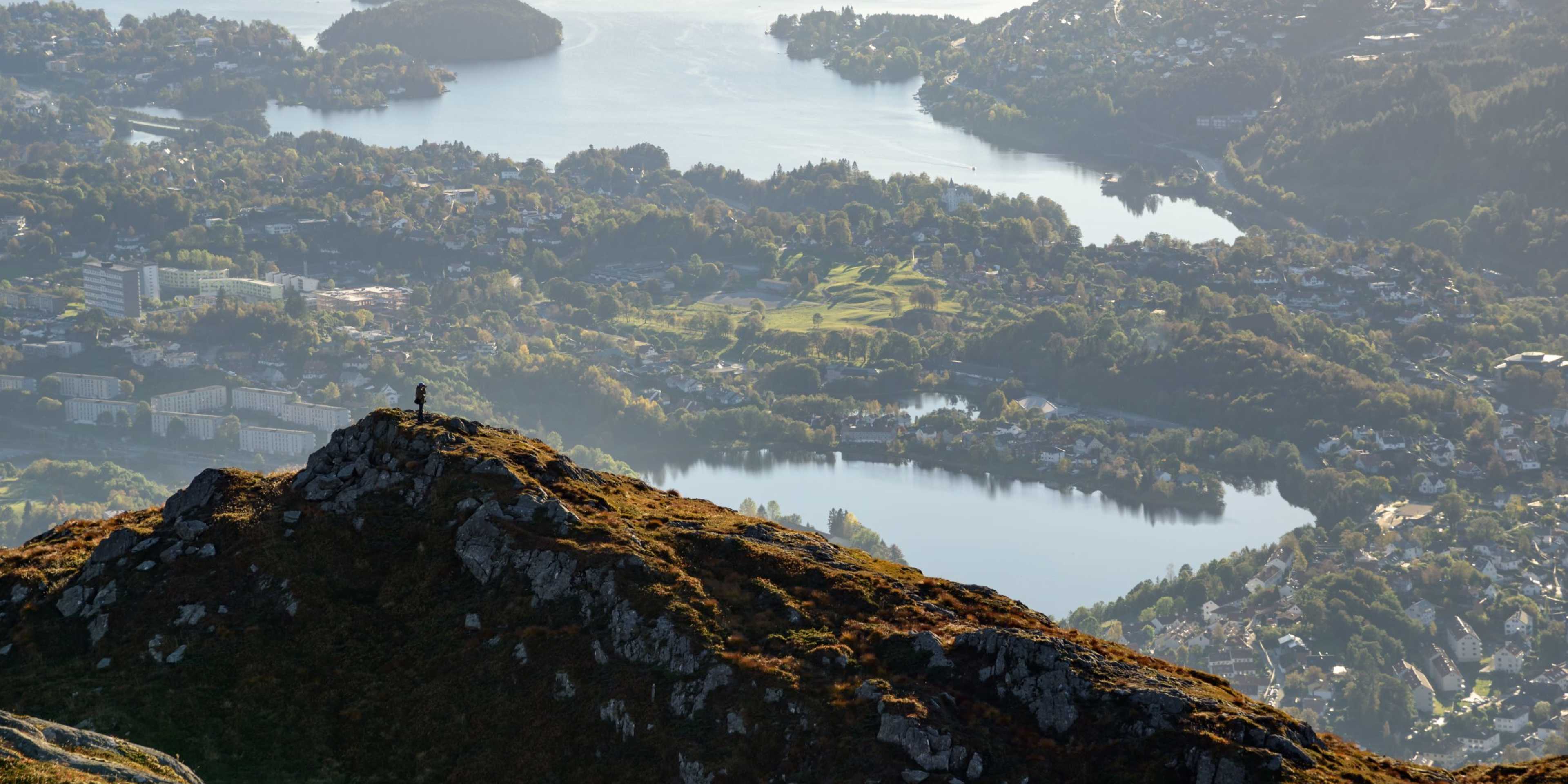 A rugged, rocky peak rises in the foreground, overlooking a lush, forested landscape with a serene lake in the distance, surrounded by towering mountains shrouded in mist.