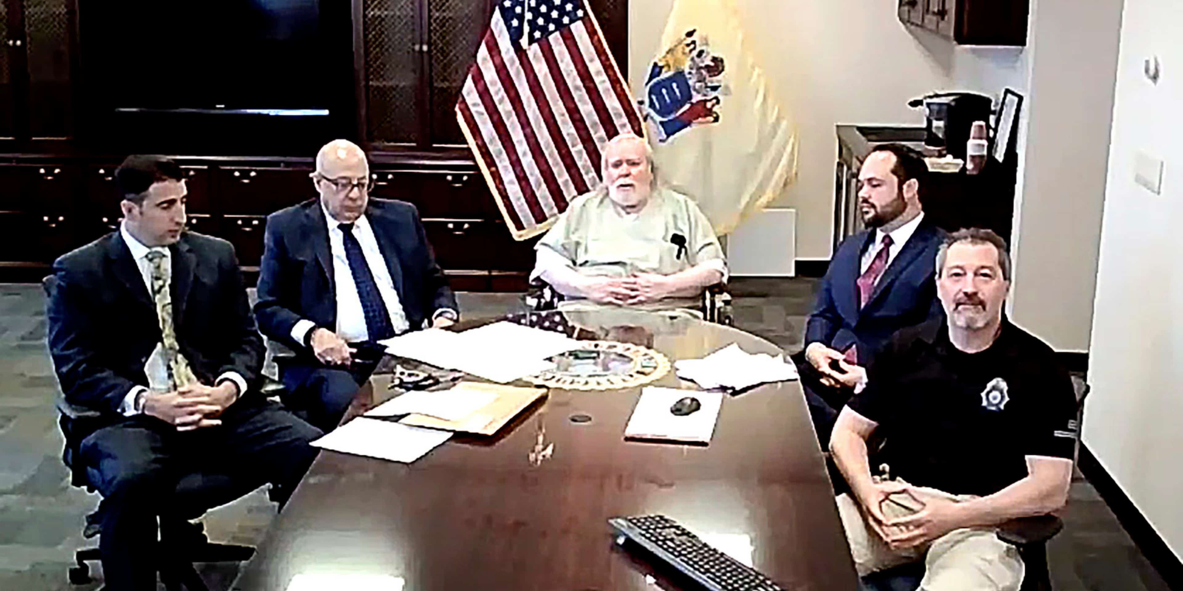 A group of men, some in suits and others in casual attire, are gathered around a table in what appears to be an office or government setting, with an American flag visible in the background.