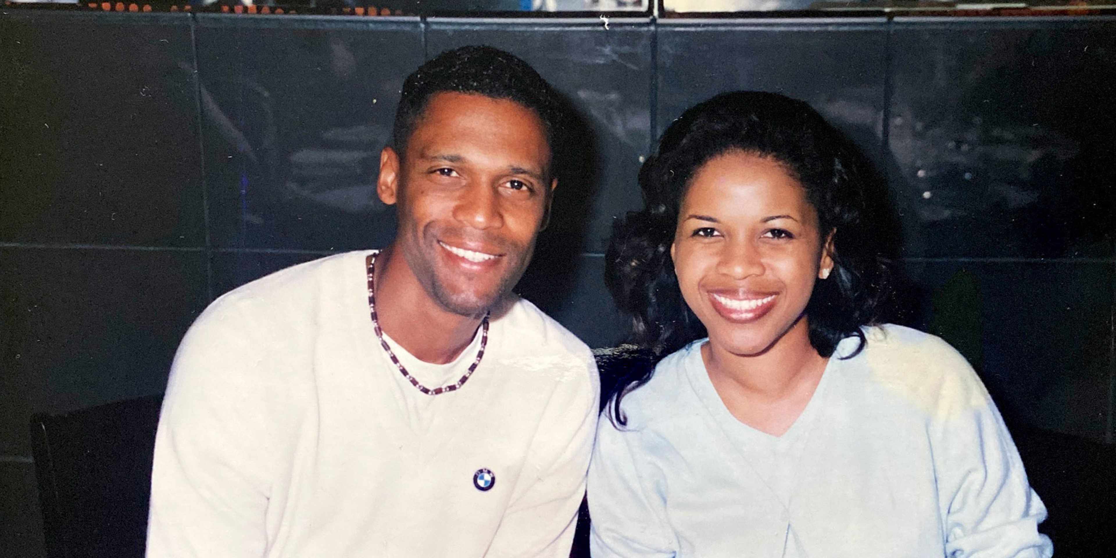 The image shows two smiling individuals, a man and a woman, standing together in what appears to be a studio or performance setting, with a dark background behind them.