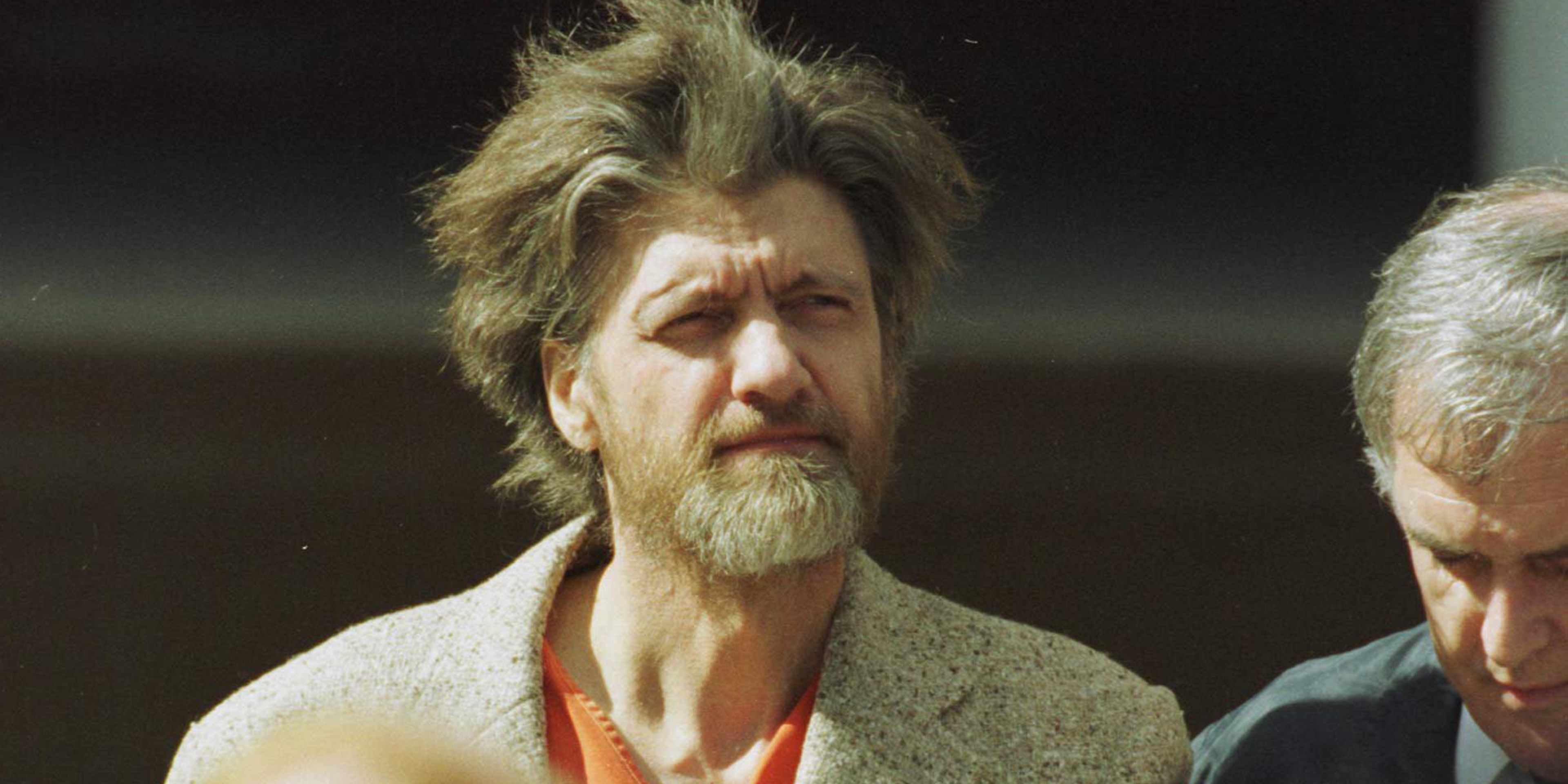 A man with a long, unkempt beard and disheveled hair stands in the foreground, while another man stands beside him in the background.