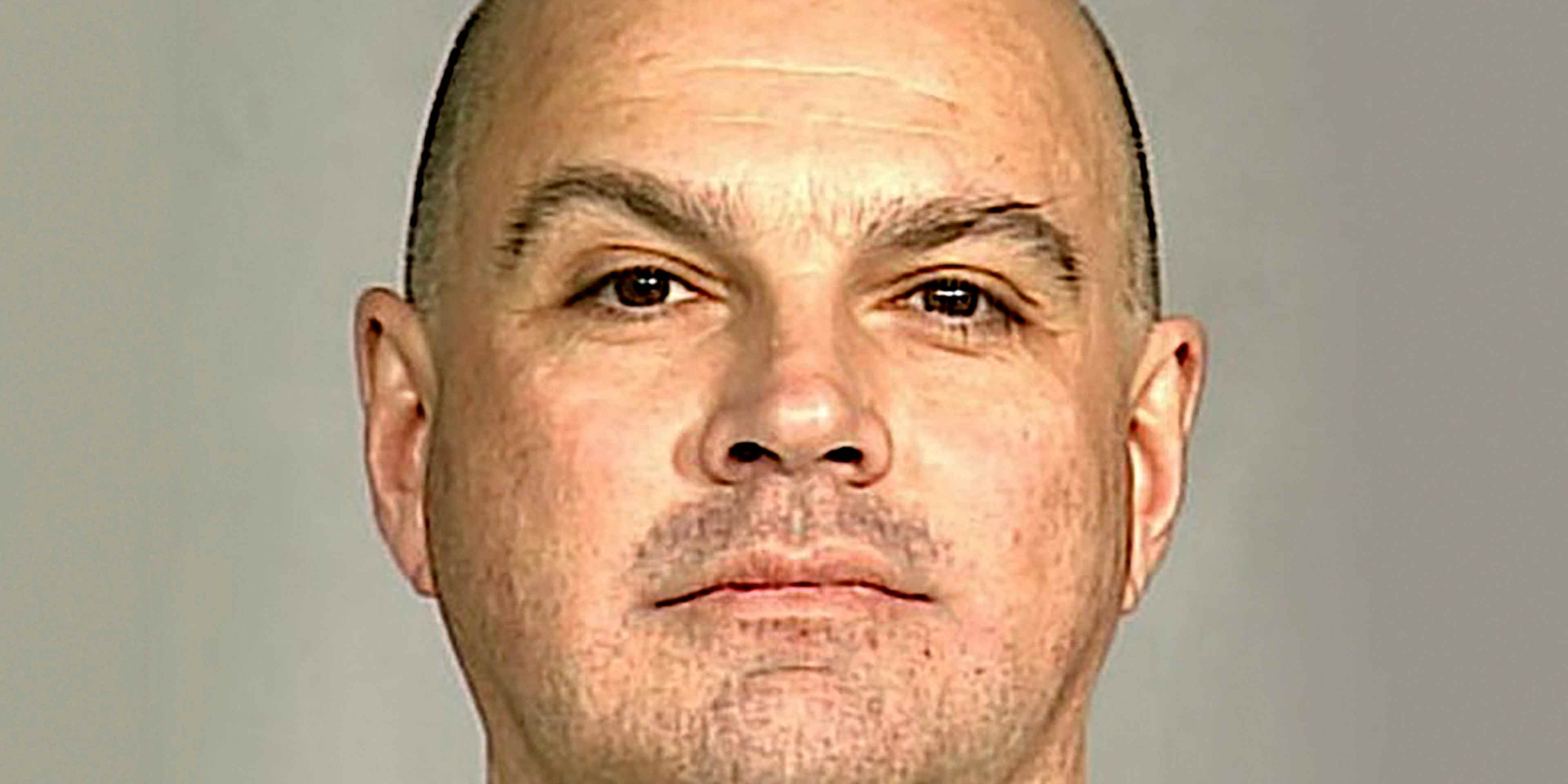 The image shows a close-up portrait of a bald, middle-aged man with a serious expression on his face, set against a plain, light-colored background.
