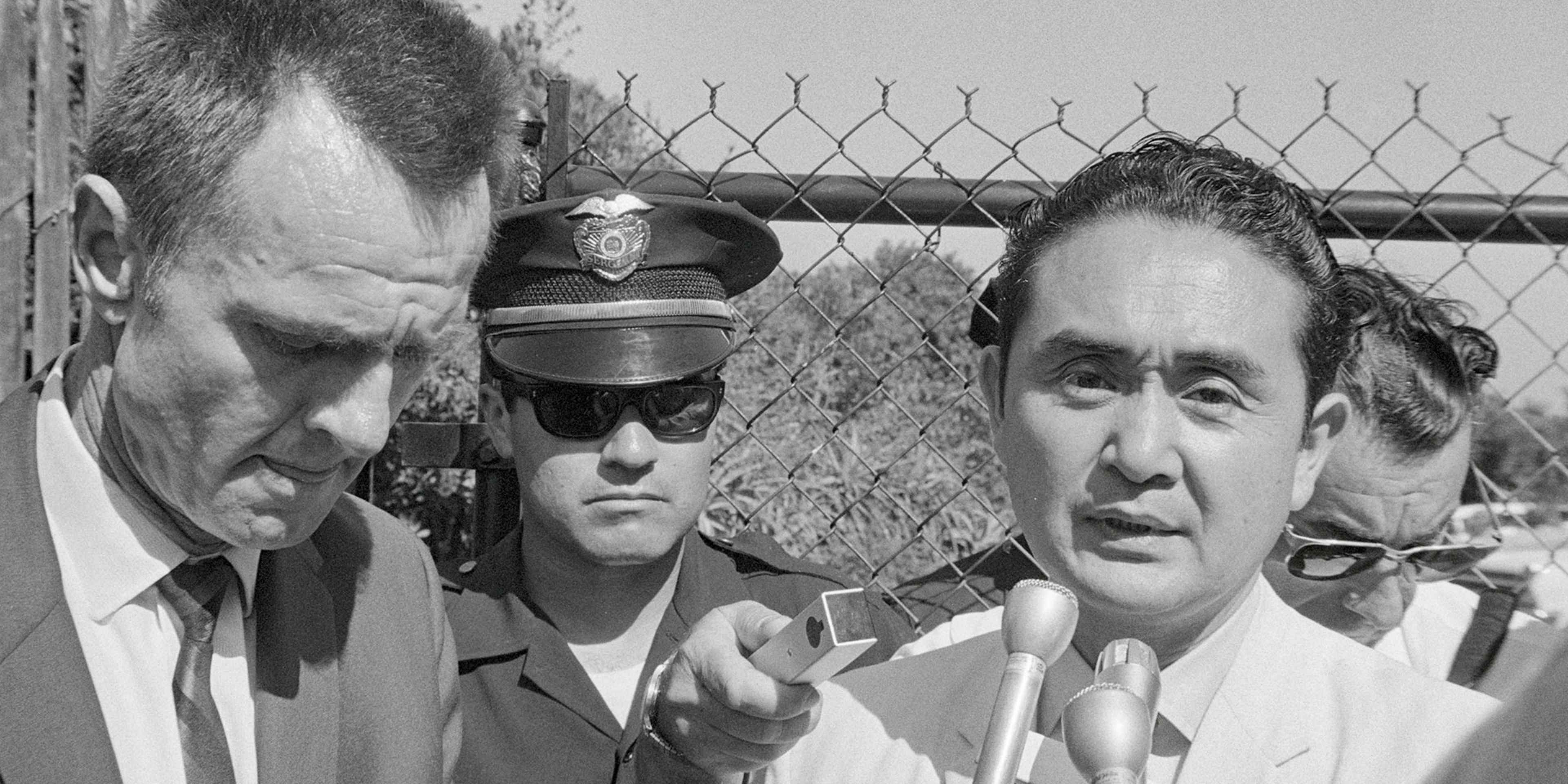 The image depicts three men, one wearing a police uniform, standing in front of a wire fence with barbed wire at the top.