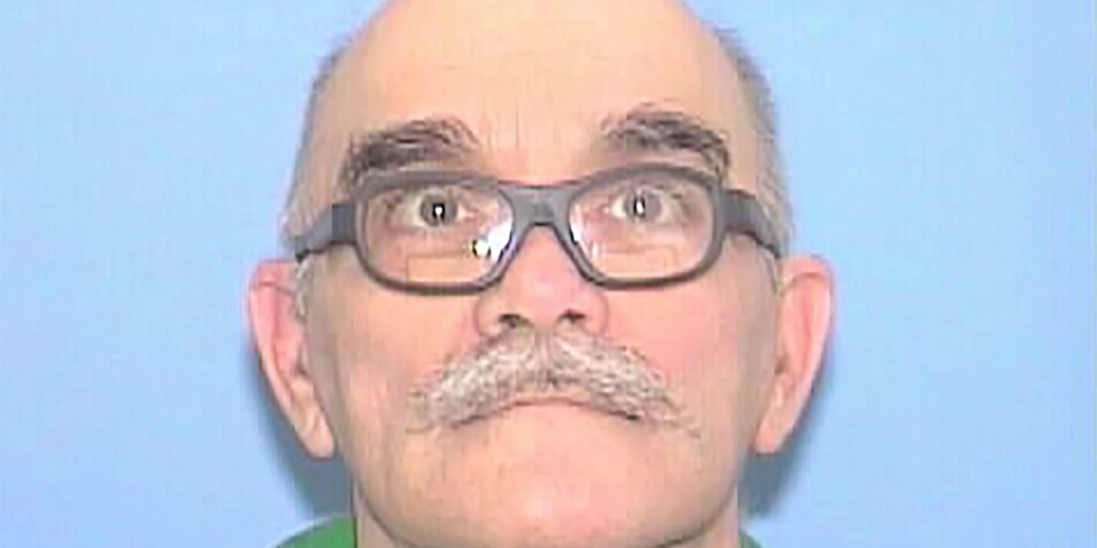 A close-up portrait of a middle-aged man with a mustache and glasses against a plain blue background.