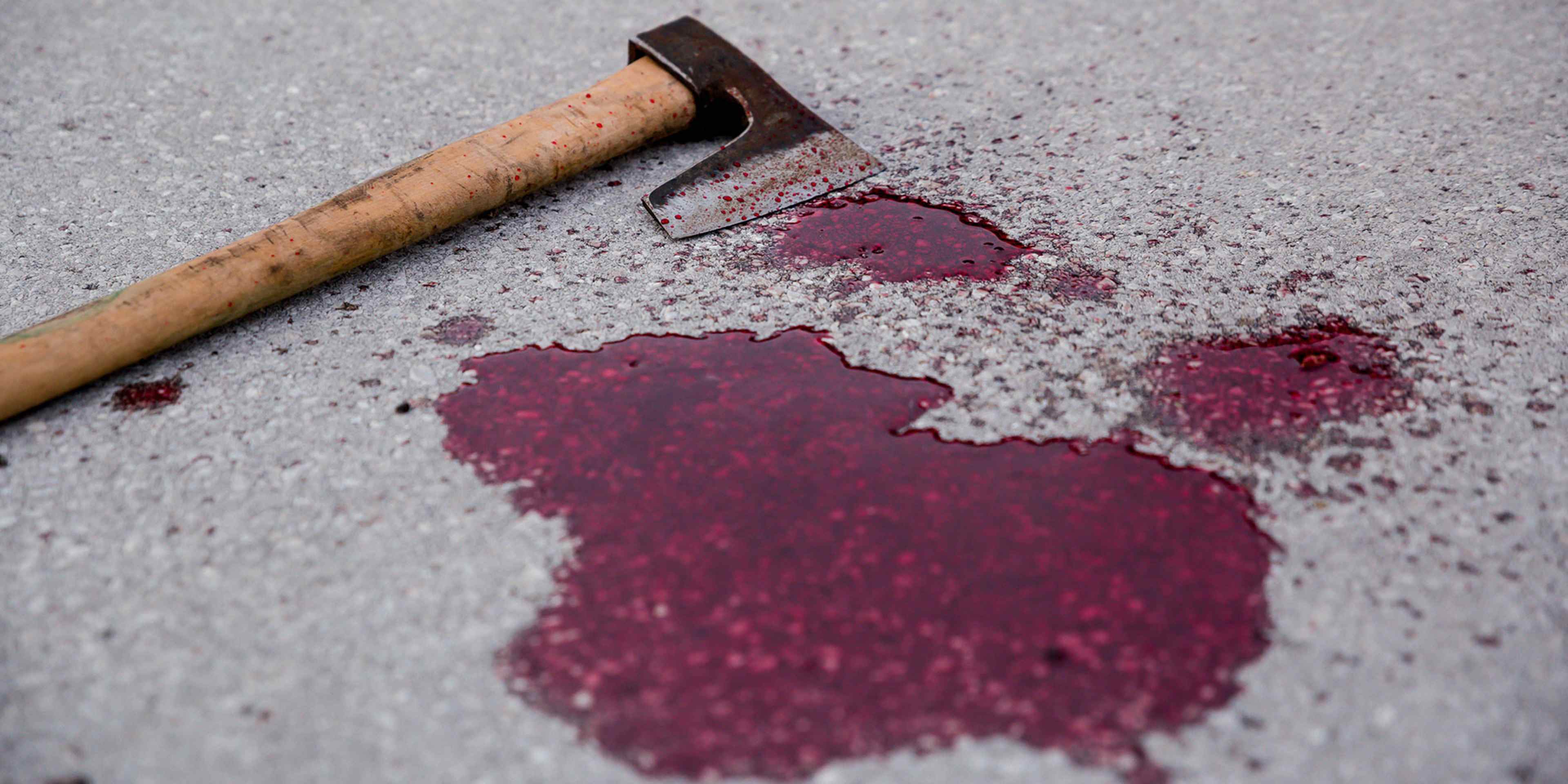 A wooden hammer lies on a concrete surface, with splatters of a deep red substance, possibly paint or blood, surrounding it.
