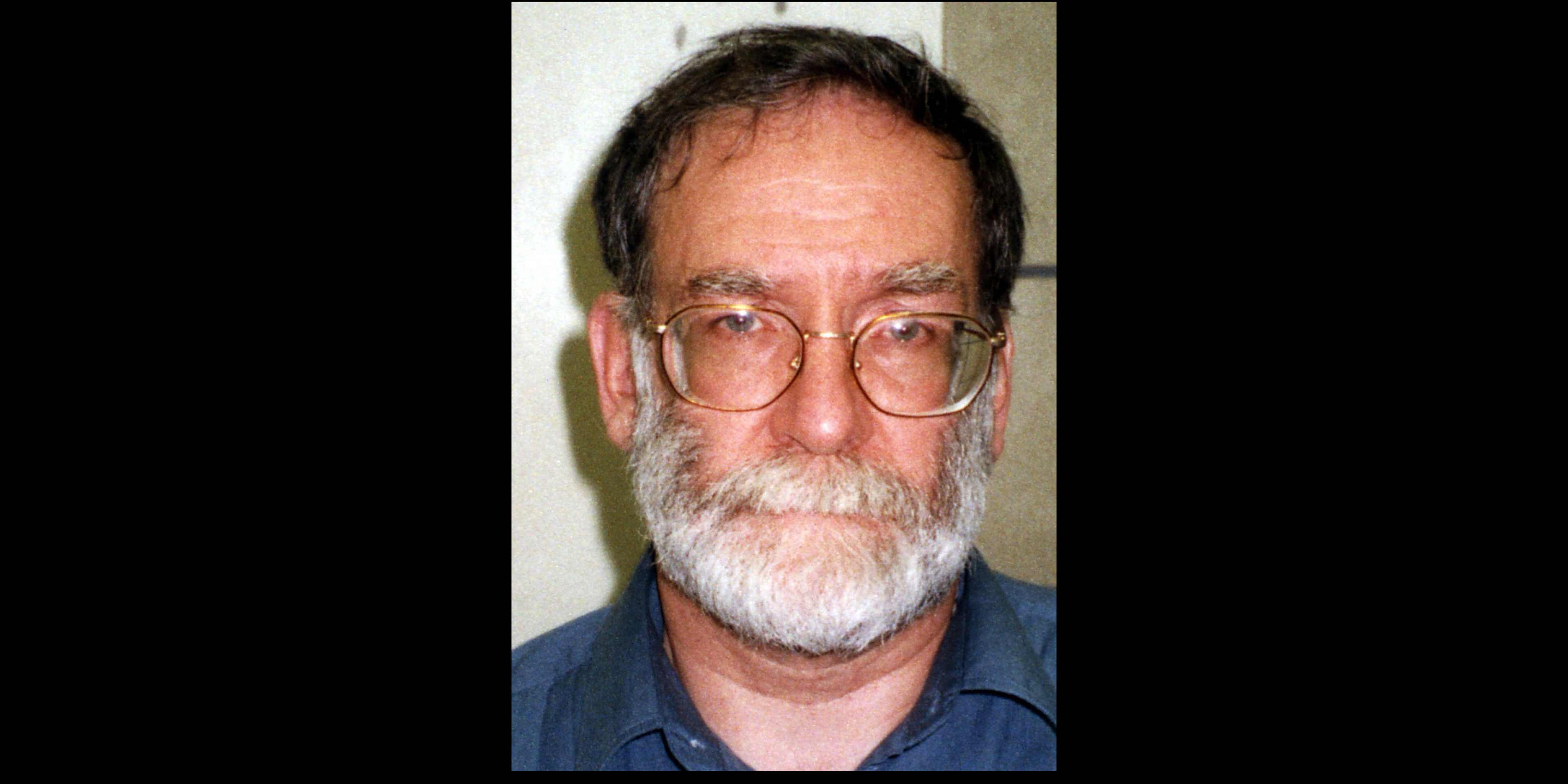 The image shows a middle-aged man with a beard and glasses, wearing a blue shirt, against a plain background.
