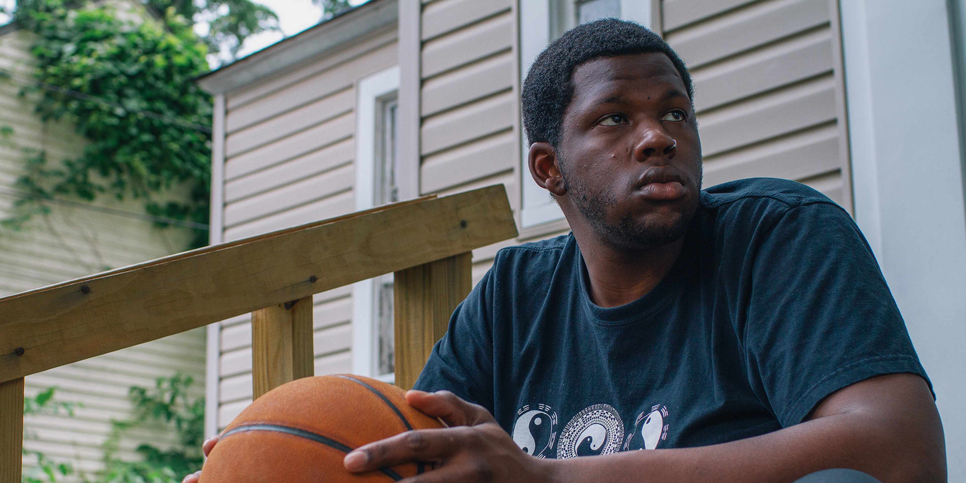 A young African American man sits on a wooden porch, holding a basketball and looking thoughtfully into the distance, with a residential building and lush greenery visible in the background.