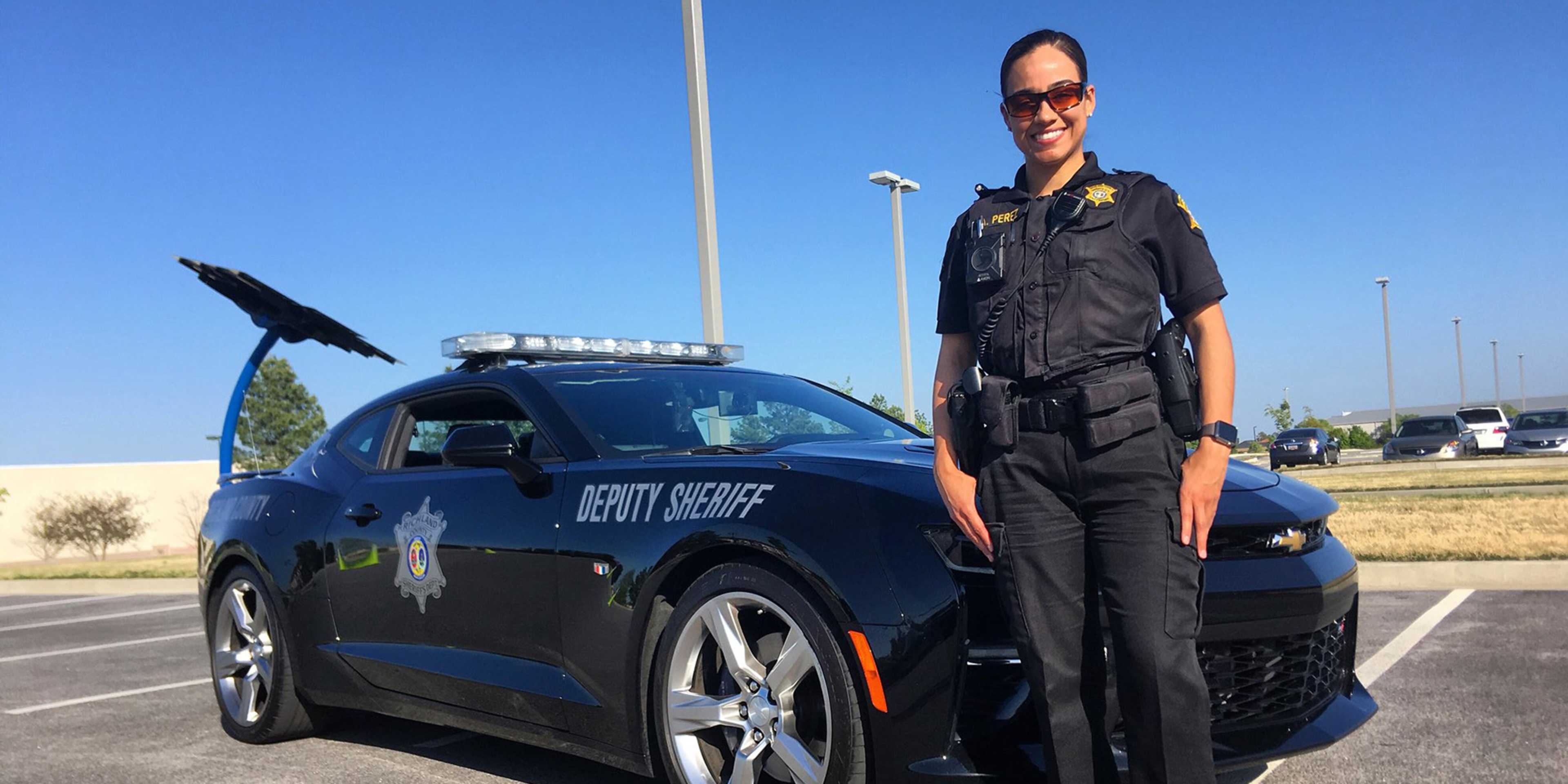 A uniformed law enforcement officer stands next to a marked police vehicle in an outdoor parking lot setting.
