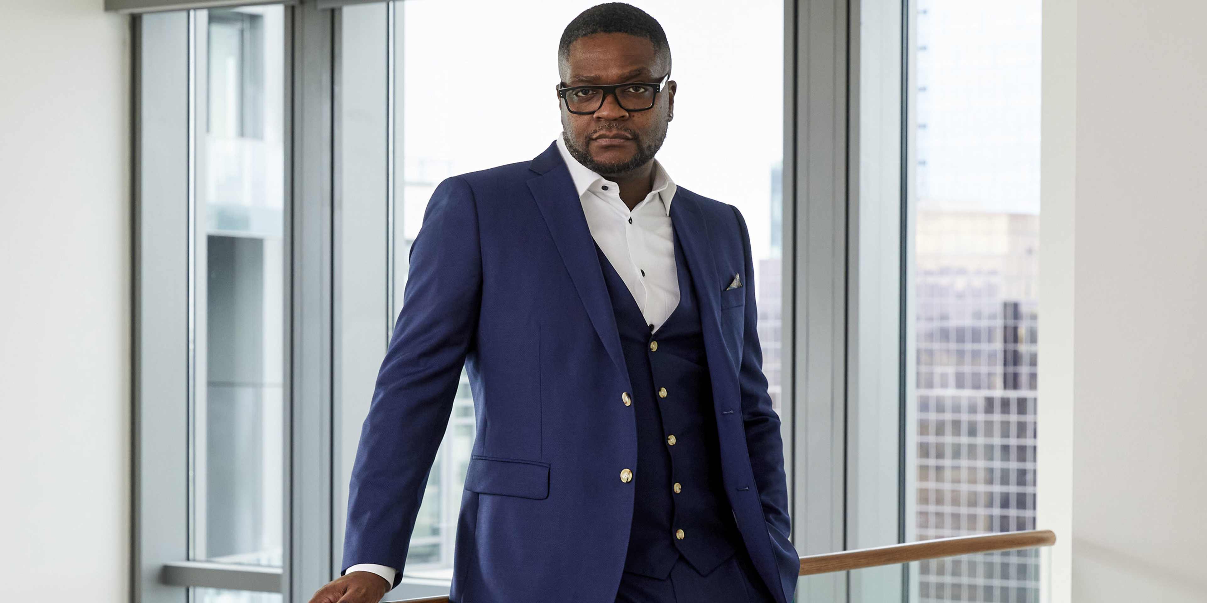 A well-dressed man in a navy blue suit stands in front of a large window, with a cityscape visible in the background.