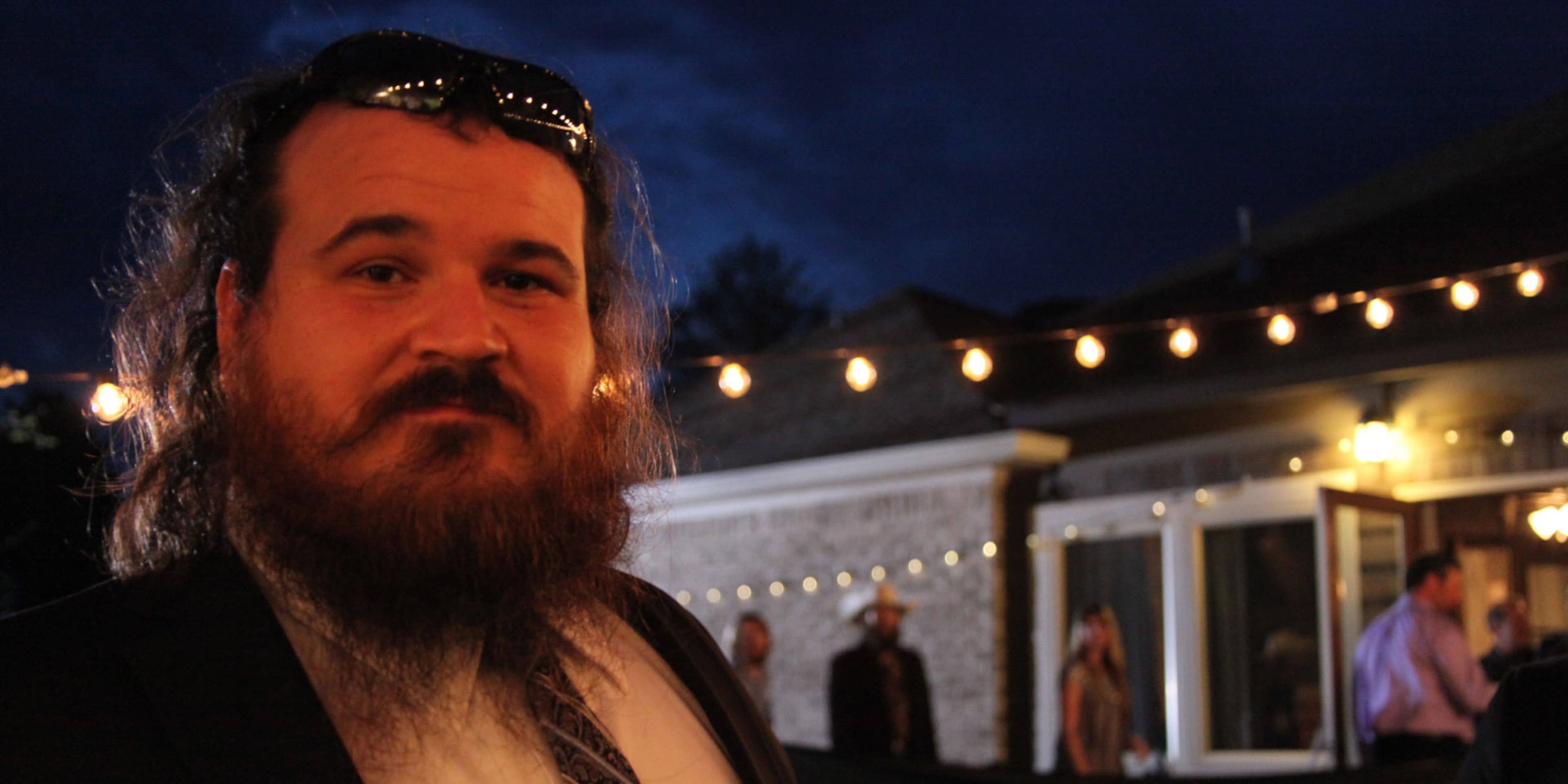 A man with a long beard and long hair stands in the foreground, surrounded by a nighttime scene with string lights and other people in the background.