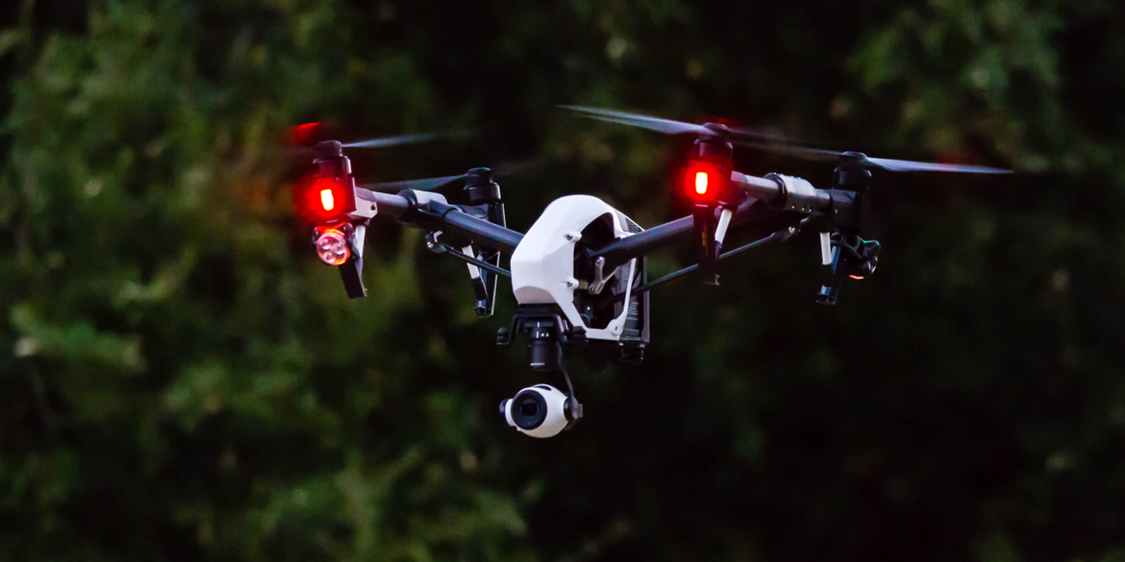 A small drone with red lights hovers in a dark, lush green environment.