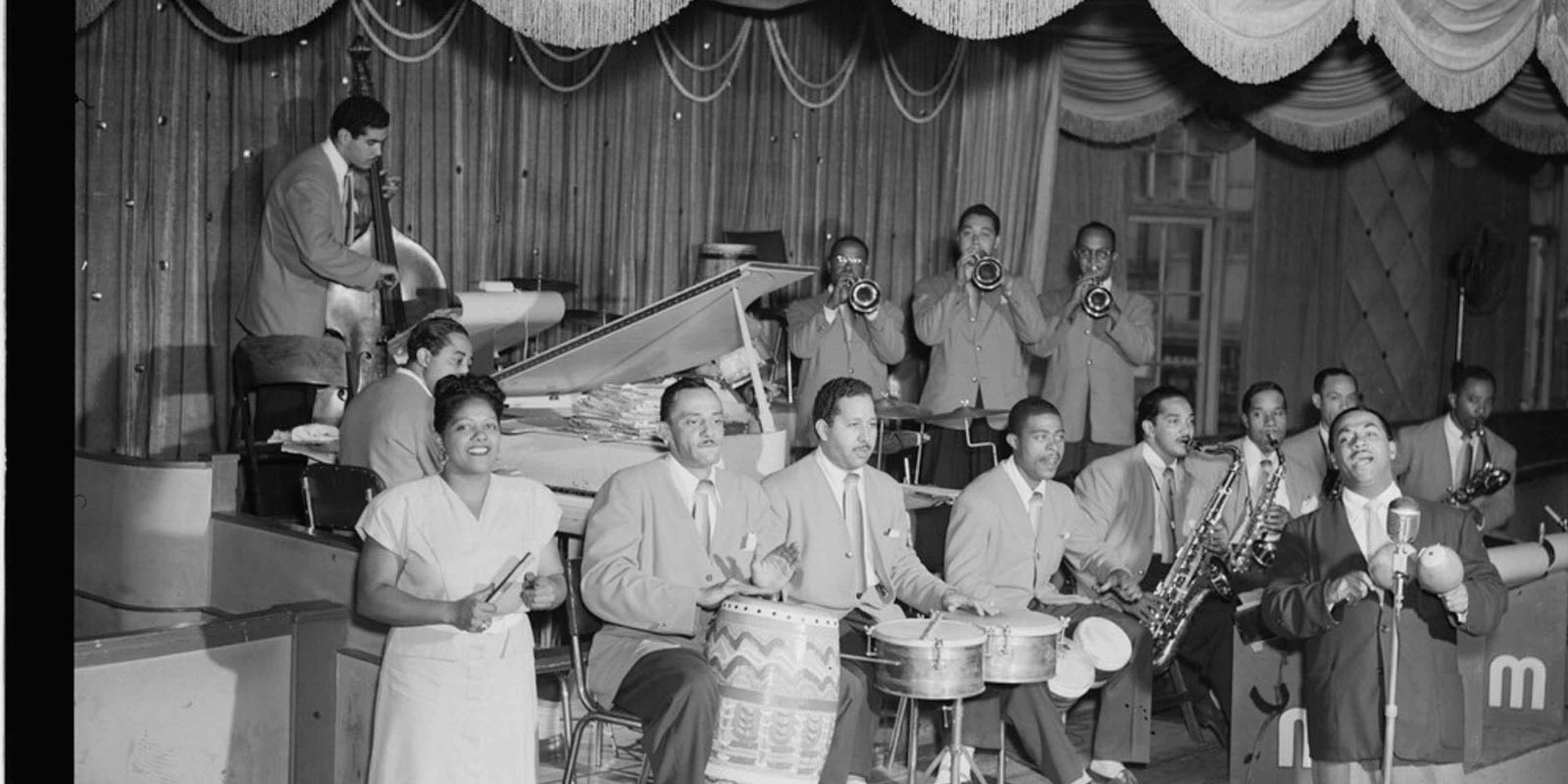 Portrait of Machito (at mic, right), with his Afro-Cuban mambo orchestra, including: José Mangual (bongos), Carlos Vidal Bolado (conga), Mario Bauzá (far left trumpeter), Ubaldo Nieto (timbales) and Graciella Grillo (front left), at the Glen Island Casino, New Rochelle, New York, c. July 1947