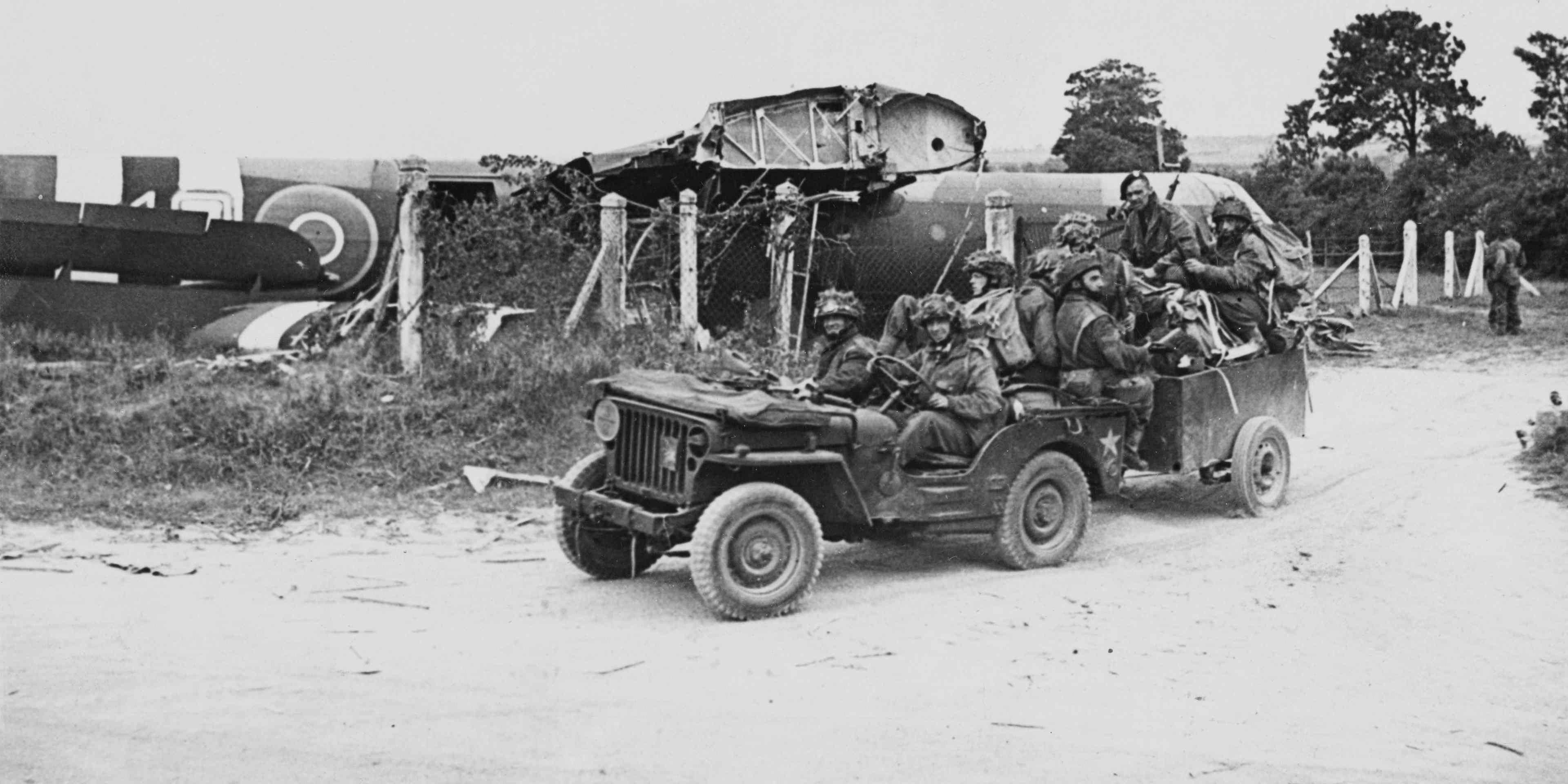 Hiram Clough (driving Jeep), Lance Corporal Joe Wilkenson and Kenneth Brierley (seated at the back of the Jeep) of the 716 (Airborne) Light Composite Company RASC (Royal Army Service Corps), British 6th Airborne Division in a Willys Jeep and trailer with glider pilots and other members of the unit following their landing from the damaged wooden Airspeed AS51 Horsa glider on a supply run on D-Day, 6th June 1944 at a landing ground near Ranville in Normandy, France.