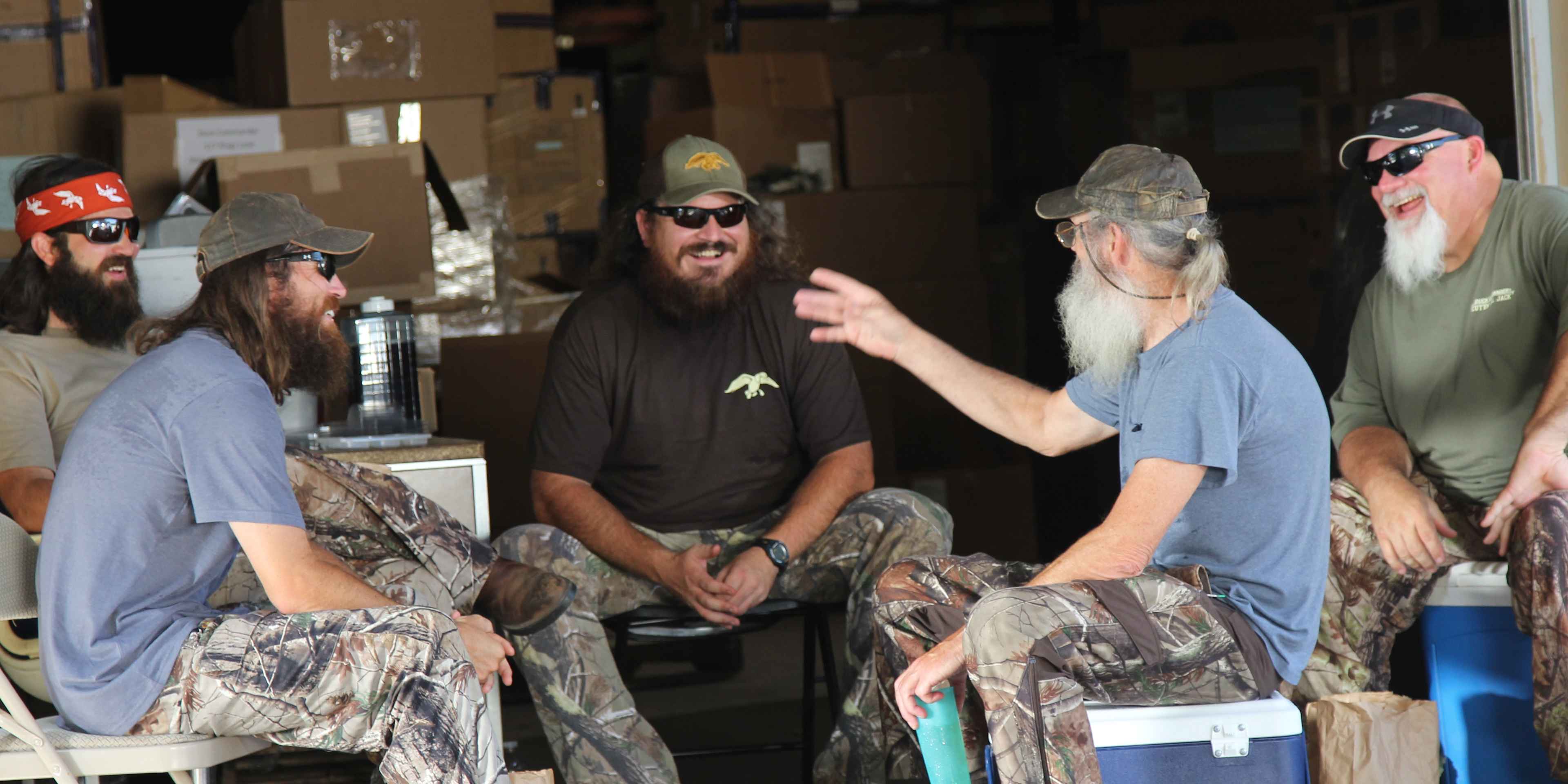 A group of men with long beards and camouflage clothing are seated in what appears to be a storage or warehouse setting, surrounded by various boxes and equipment.