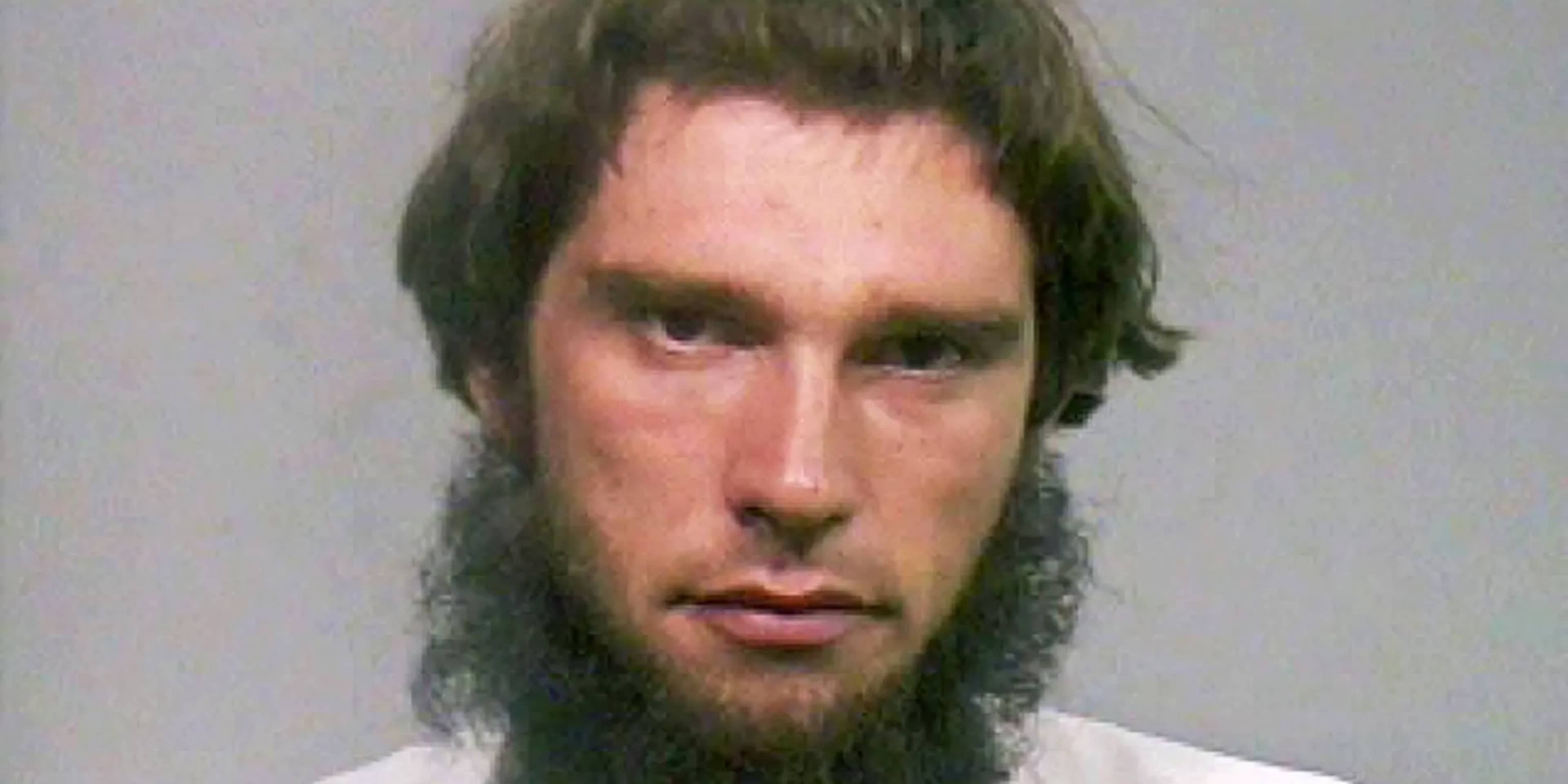 A man with long, dark hair and a beard is the focal point of the image, set against a plain, light-colored background.