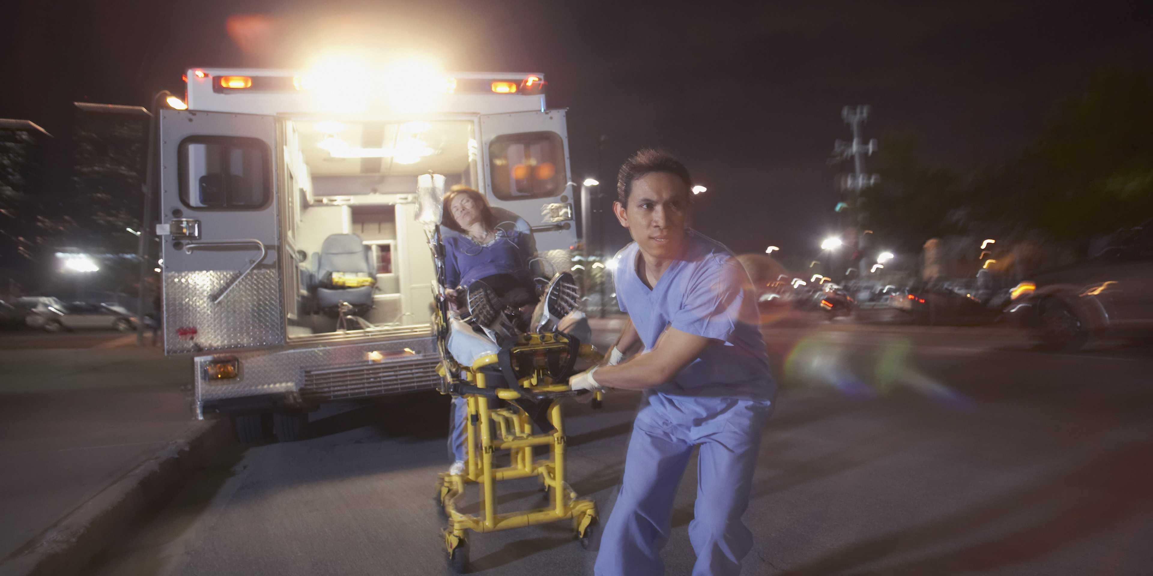 The image shows a nighttime scene with an ambulance in the background, its lights illuminating the area, and a person in medical scrubs standing in the foreground. Photo of EMS practitioner. Credit: ERproduction Ltd/Blend Images