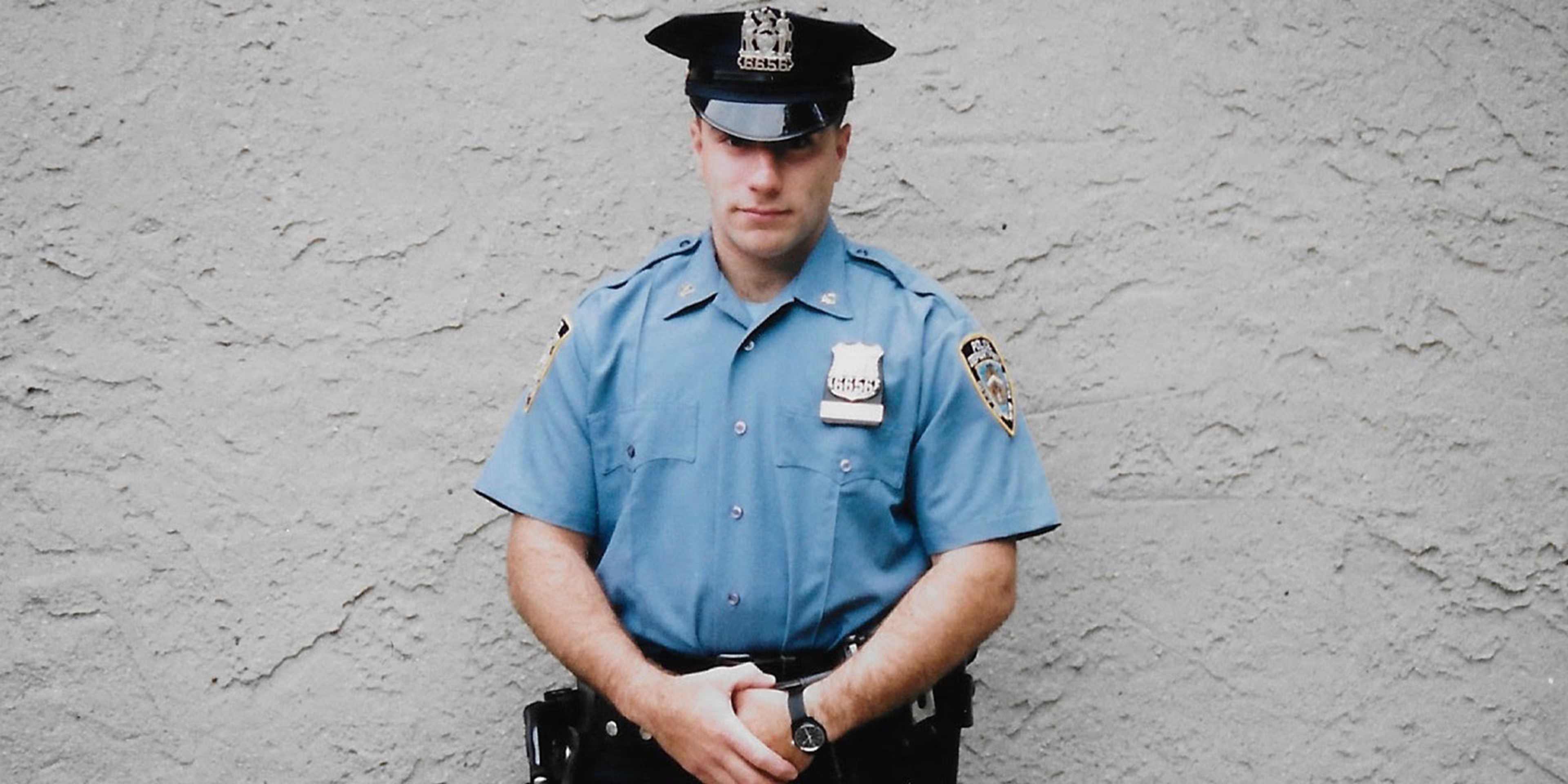 A uniformed police officer stands in front of a plain, textured wall, holding a baton and wearing a badge on their uniform.