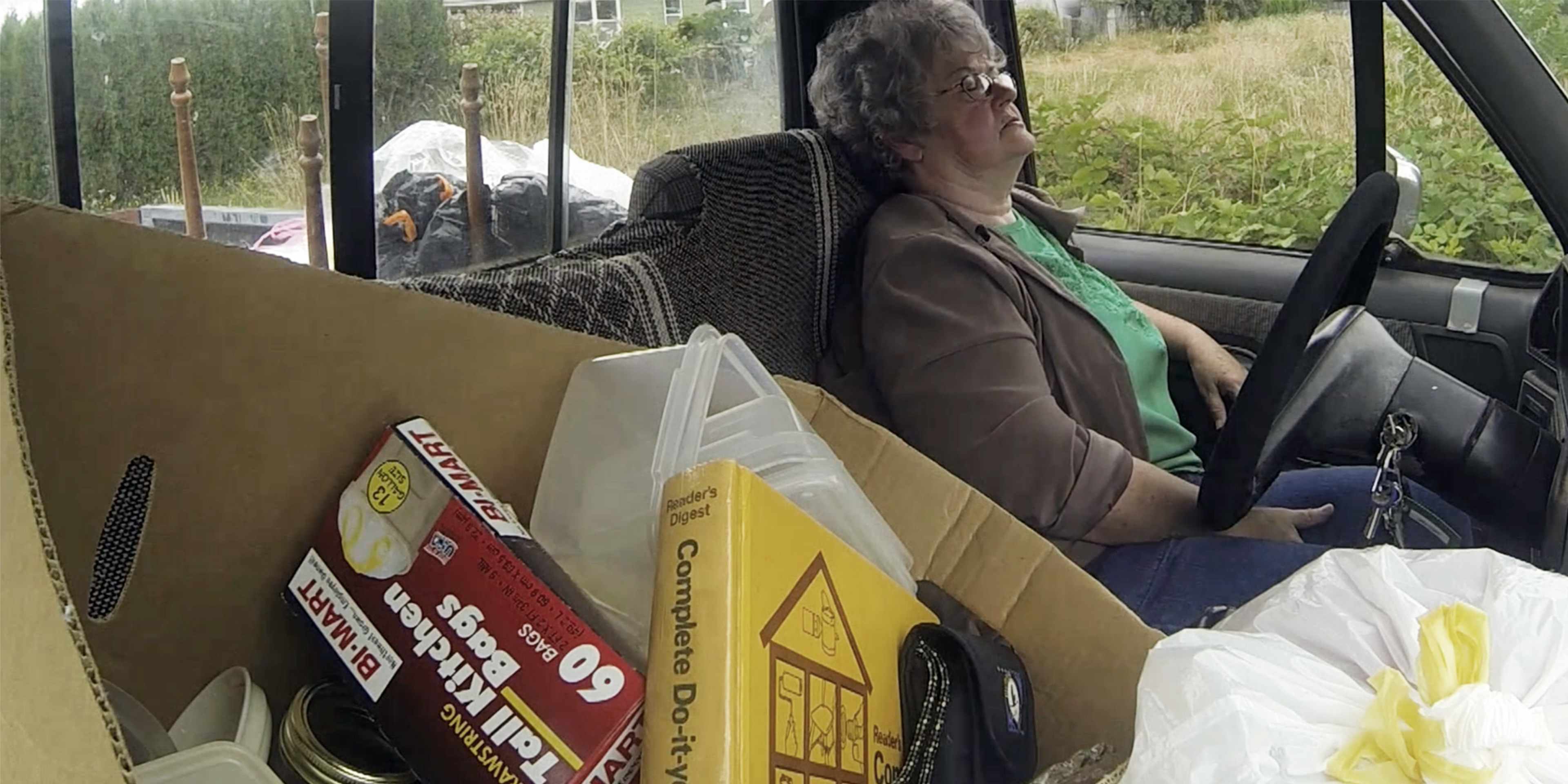 A person sits in the driver's seat of a vehicle, surrounded by various boxes and bags, suggesting they are in the process of moving or transporting items.