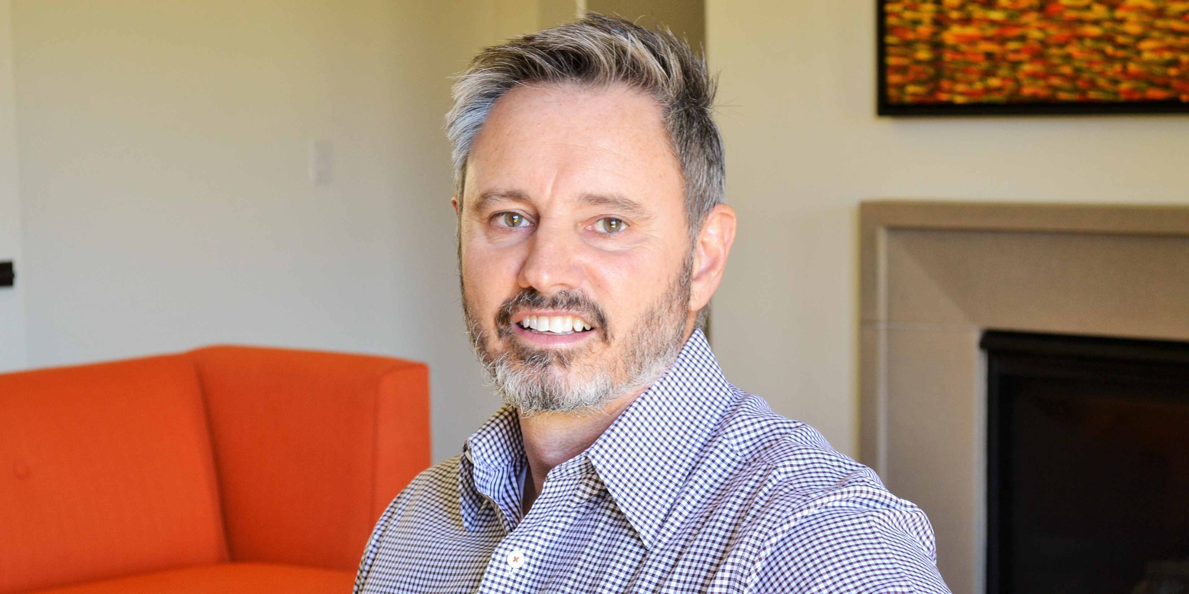 The image shows a middle-aged man with gray hair and a beard, wearing a checkered shirt, sitting on an orange couch in what appears to be a living room setting with a framed artwork on the wall behind him.
