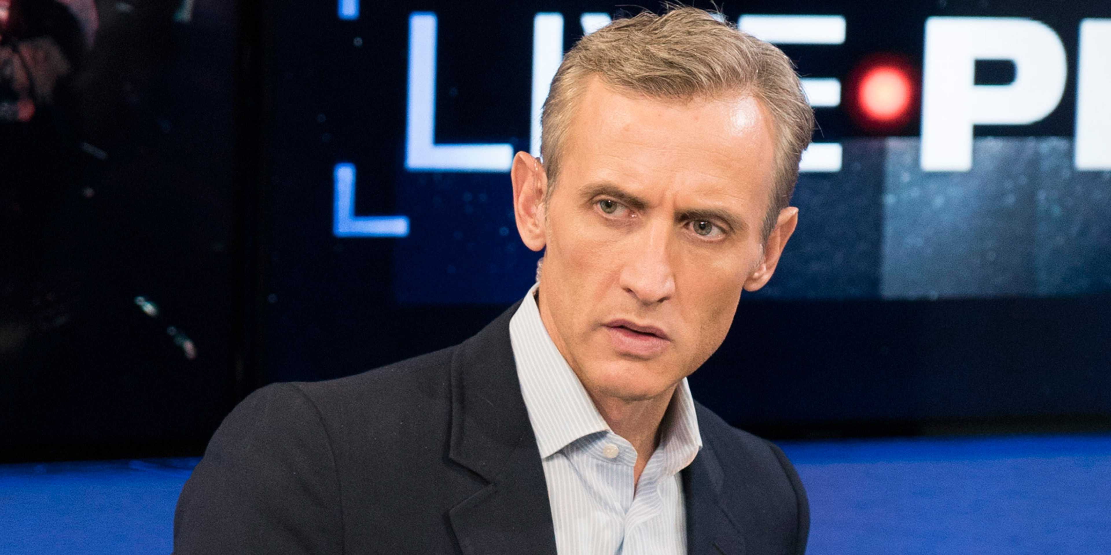 A middle-aged man with blond hair is standing in front of a blue backdrop with the word "LIVE" displayed prominently.