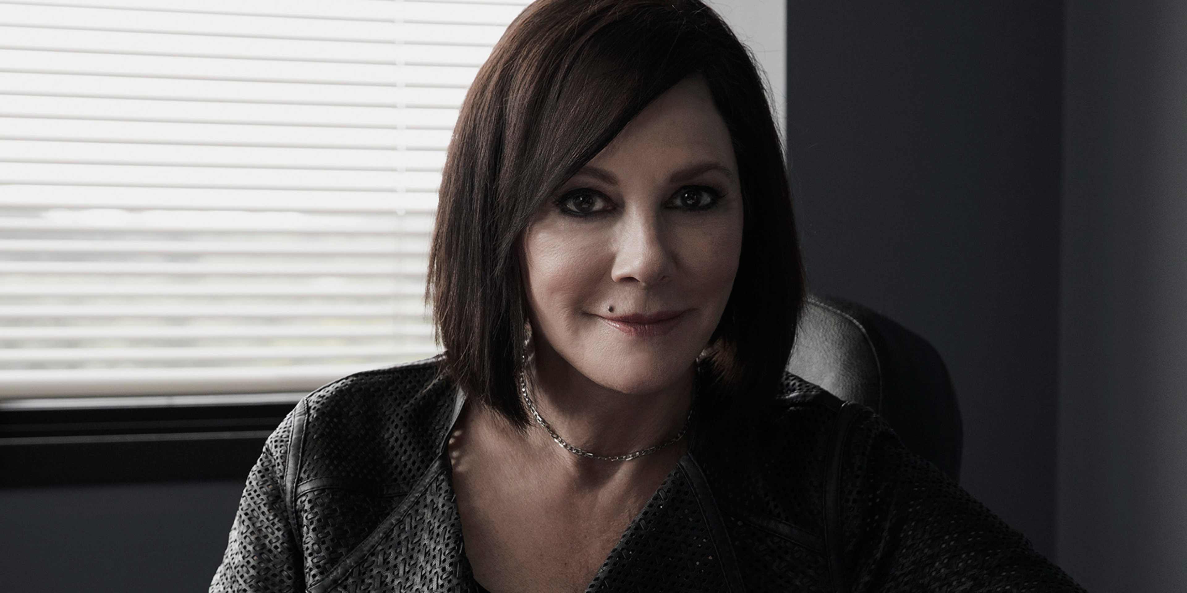 A woman with dark hair and a serious expression is sitting near a window, with a blurred background visible behind her.