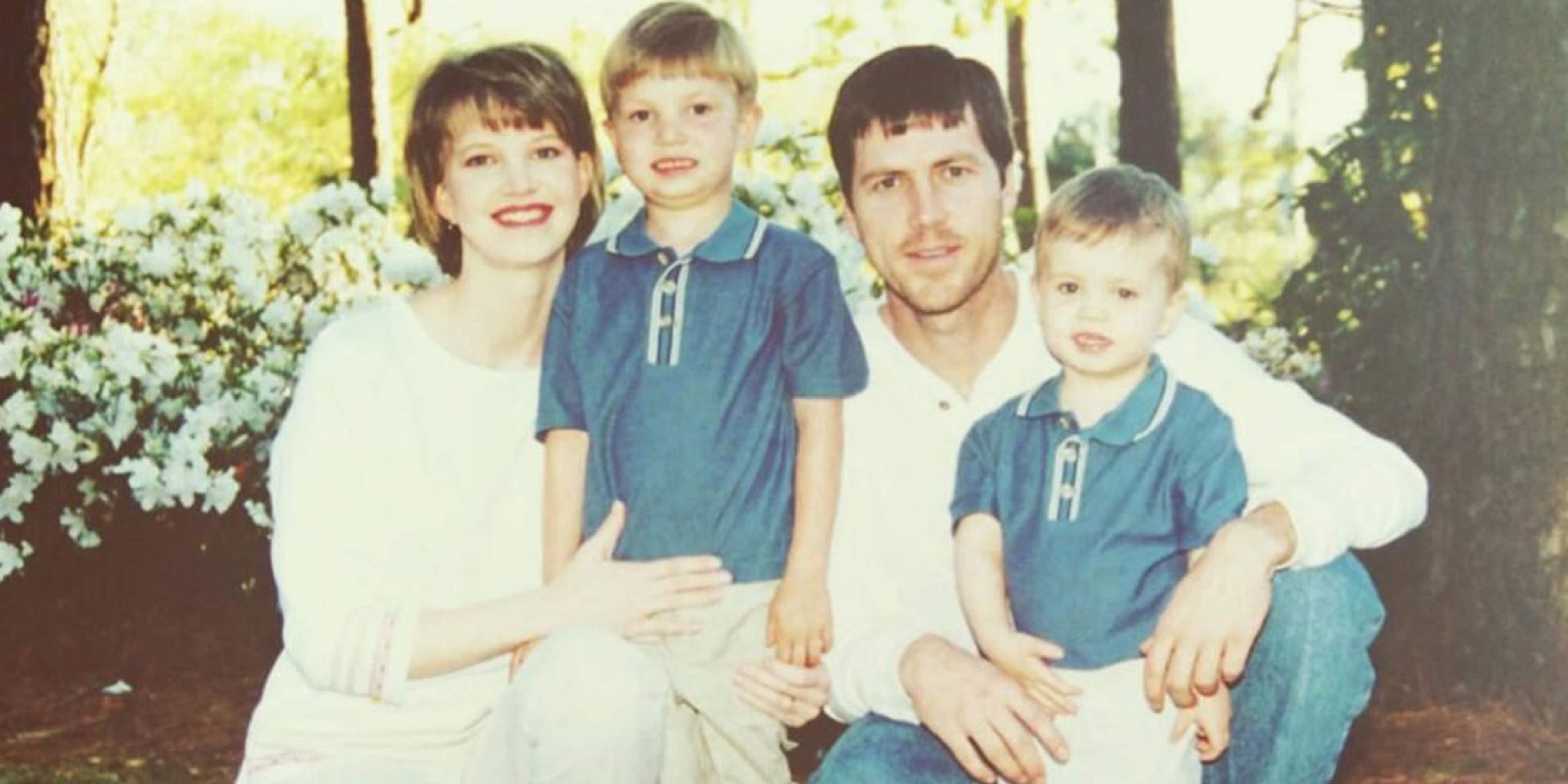 A family of four, including a mother, father, and two young children, posing together outdoors in a garden setting with blooming flowers in the background.
