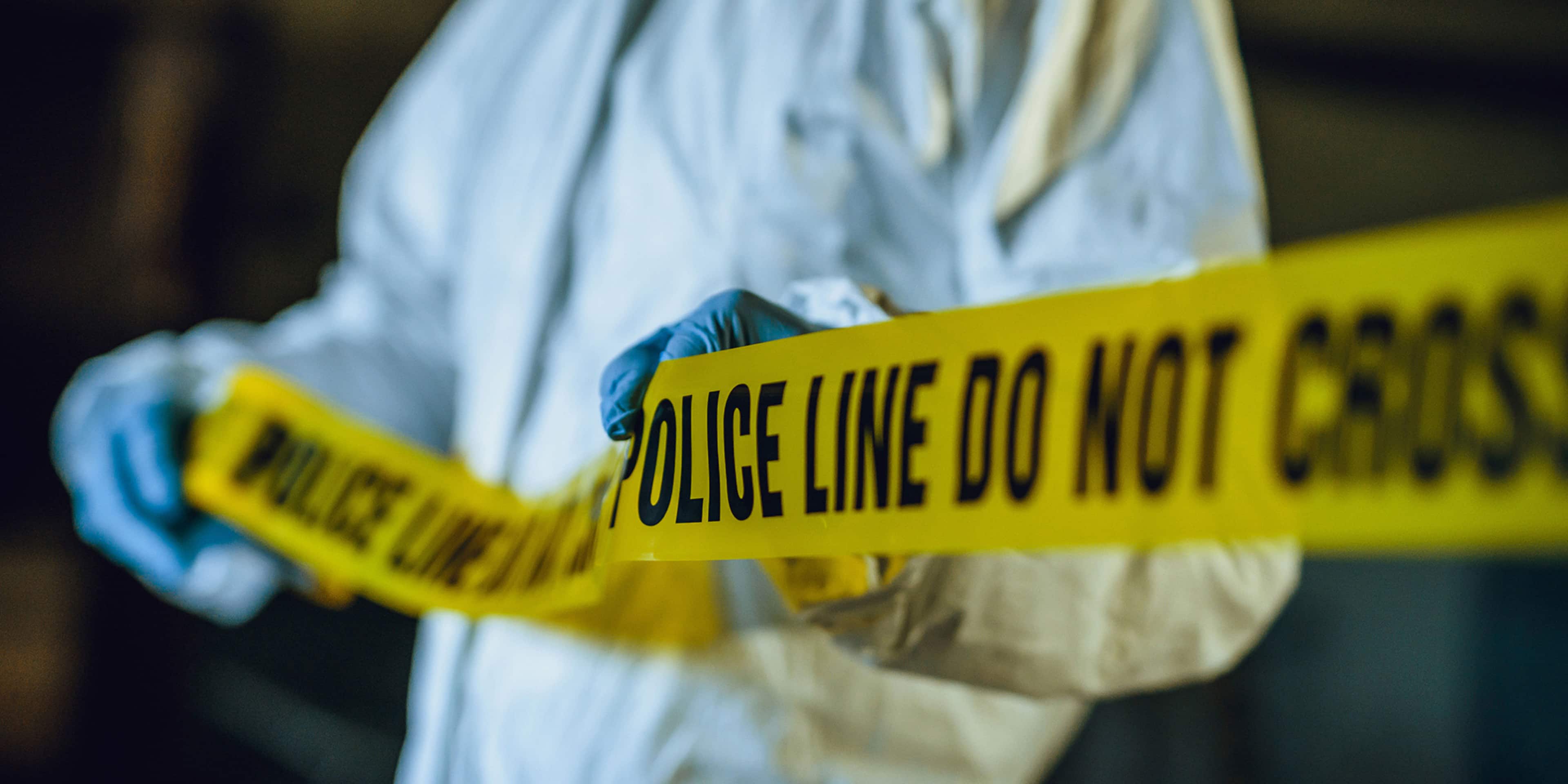 The image shows a person wearing protective gear, such as a white hazmat suit, standing behind a yellow police line that reads "POLICE LINE DO NOT CROSS".