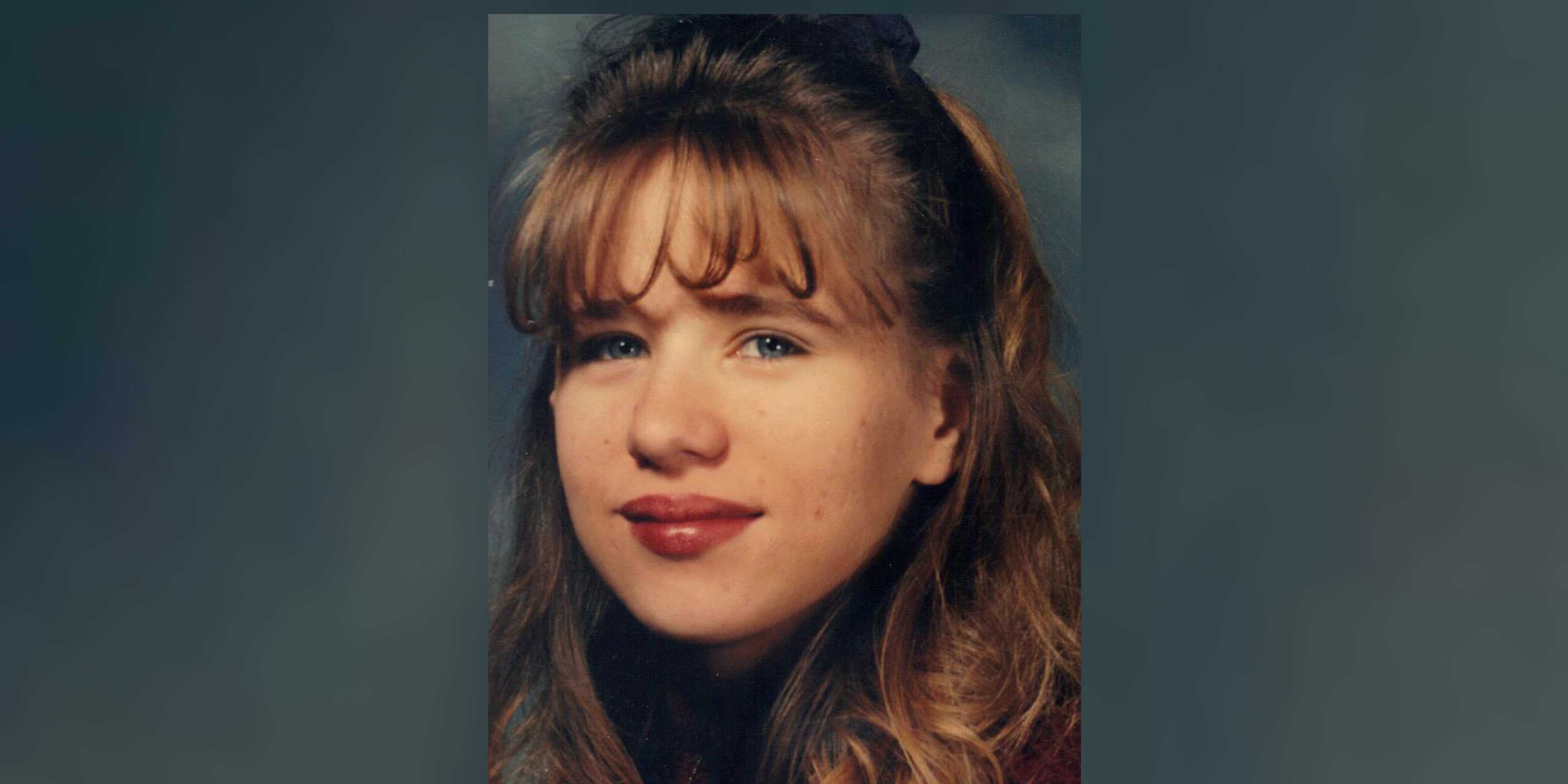 A close-up portrait of a young woman with long, wavy brown hair and a thoughtful expression on her face, set against a dark background.