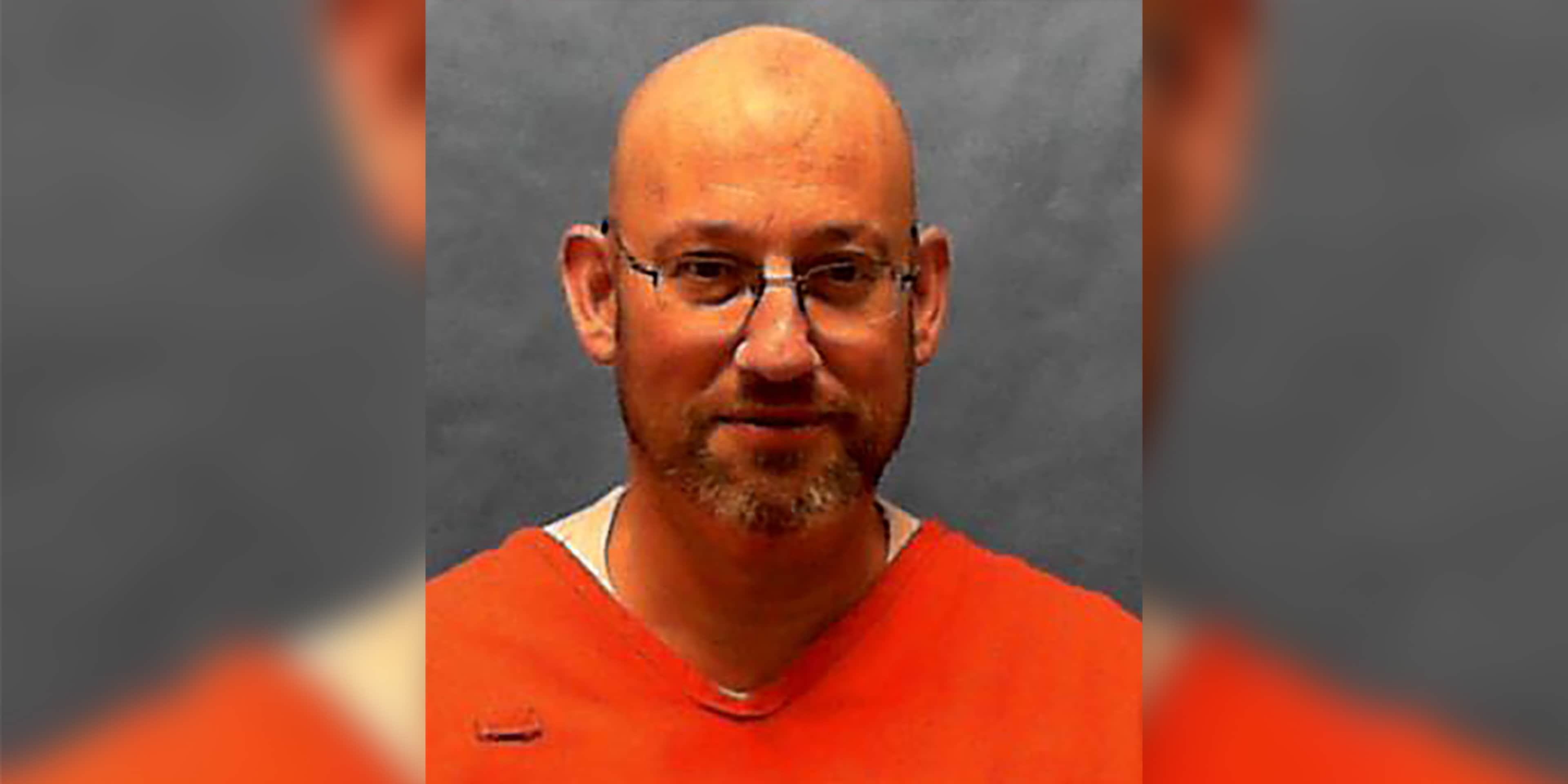 A middle-aged man with a beard and glasses, wearing an orange shirt, appears against a gray background.