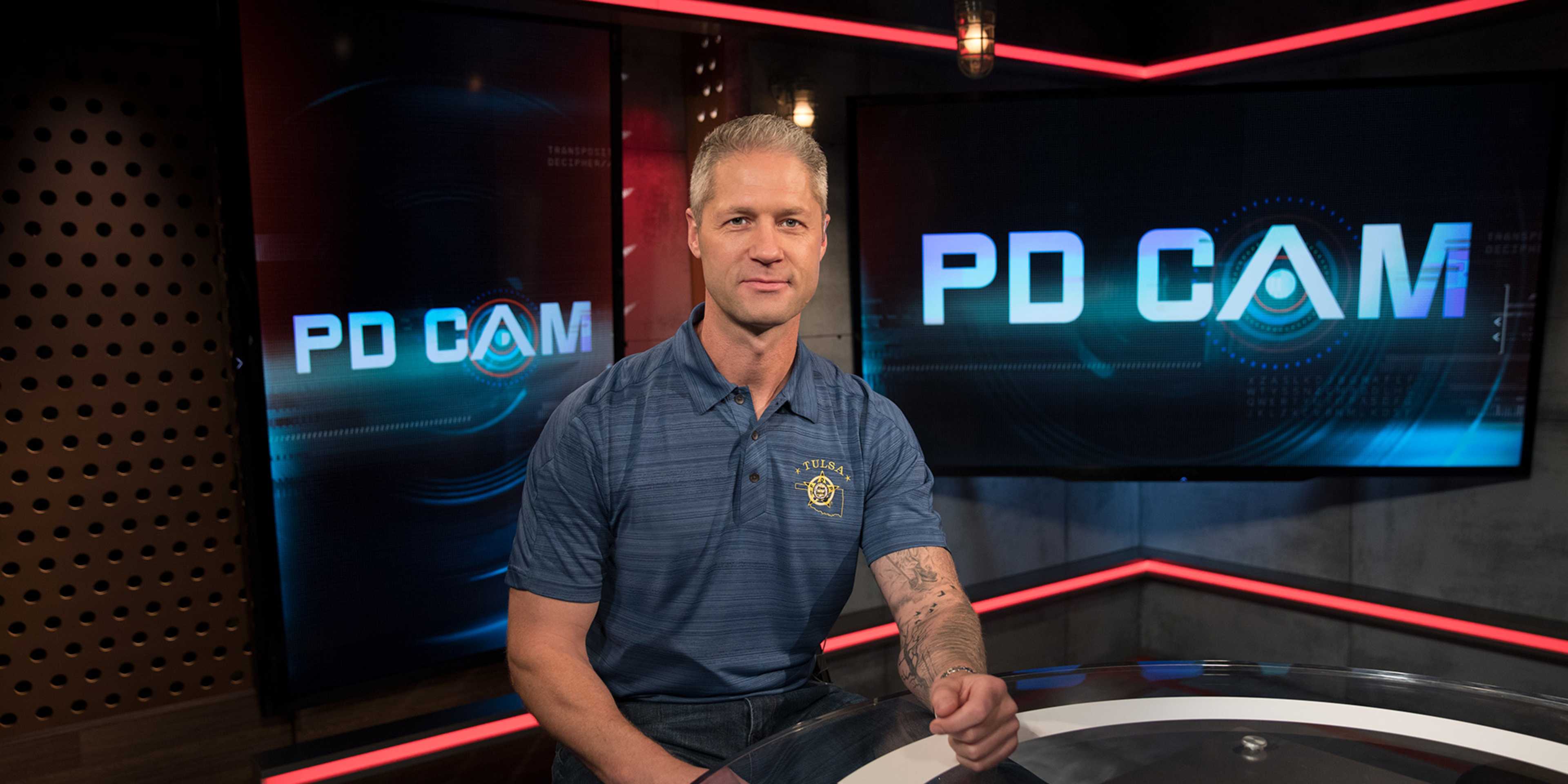 A man in a police uniform stands in front of a backdrop with the text "PD CAM" displayed prominently.
