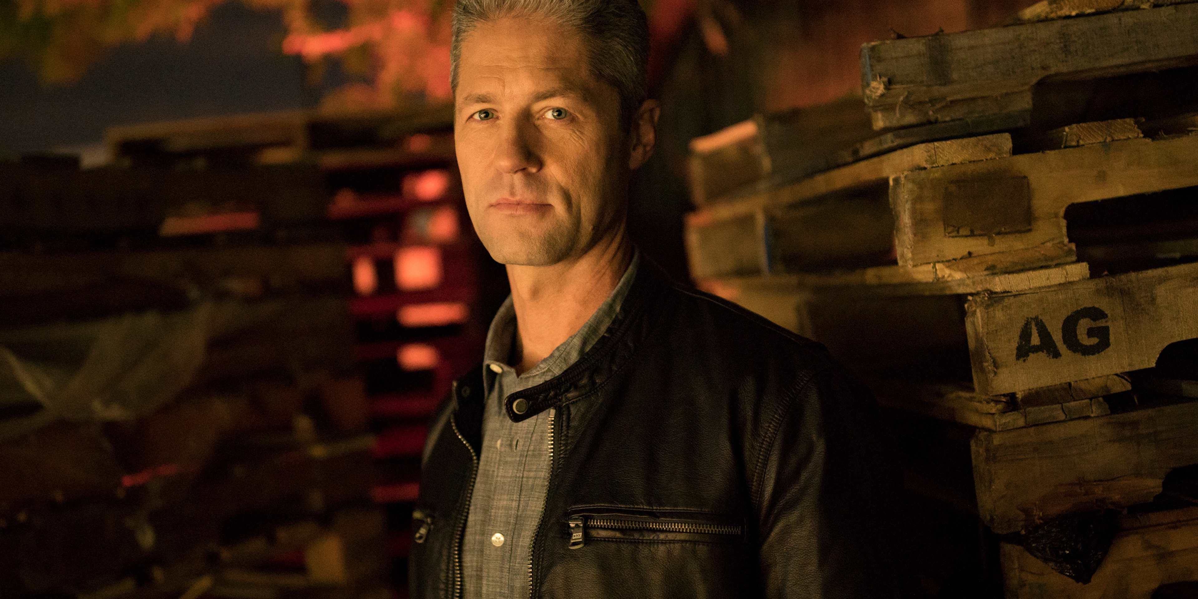 The image shows a middle-aged man with gray hair and a serious expression, standing in front of a brick wall with a wooden crate labeled "AG" in the background.