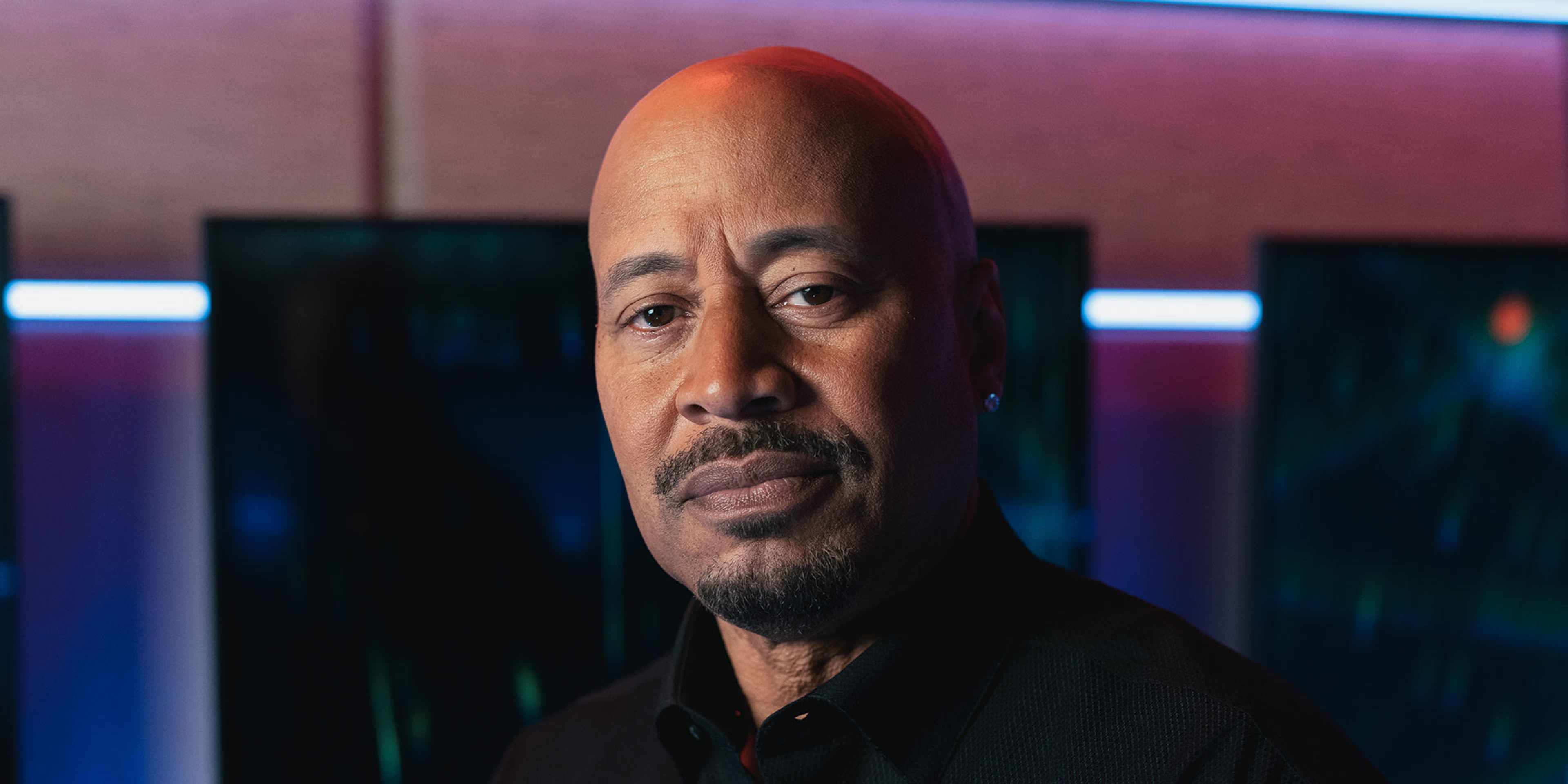 The image shows a middle-aged man with a serious expression, wearing a black jacket, against a colorful, abstract background.