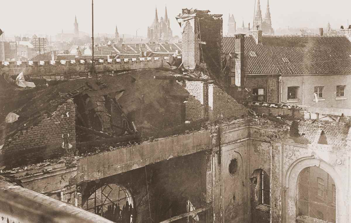 A sepia-toned photograph depicting a ruined cityscape with damaged buildings, rubble, and a partially collapsed archway in the foreground, while in the background, the spires of a cathedral or church can be seen rising above the rooftops.