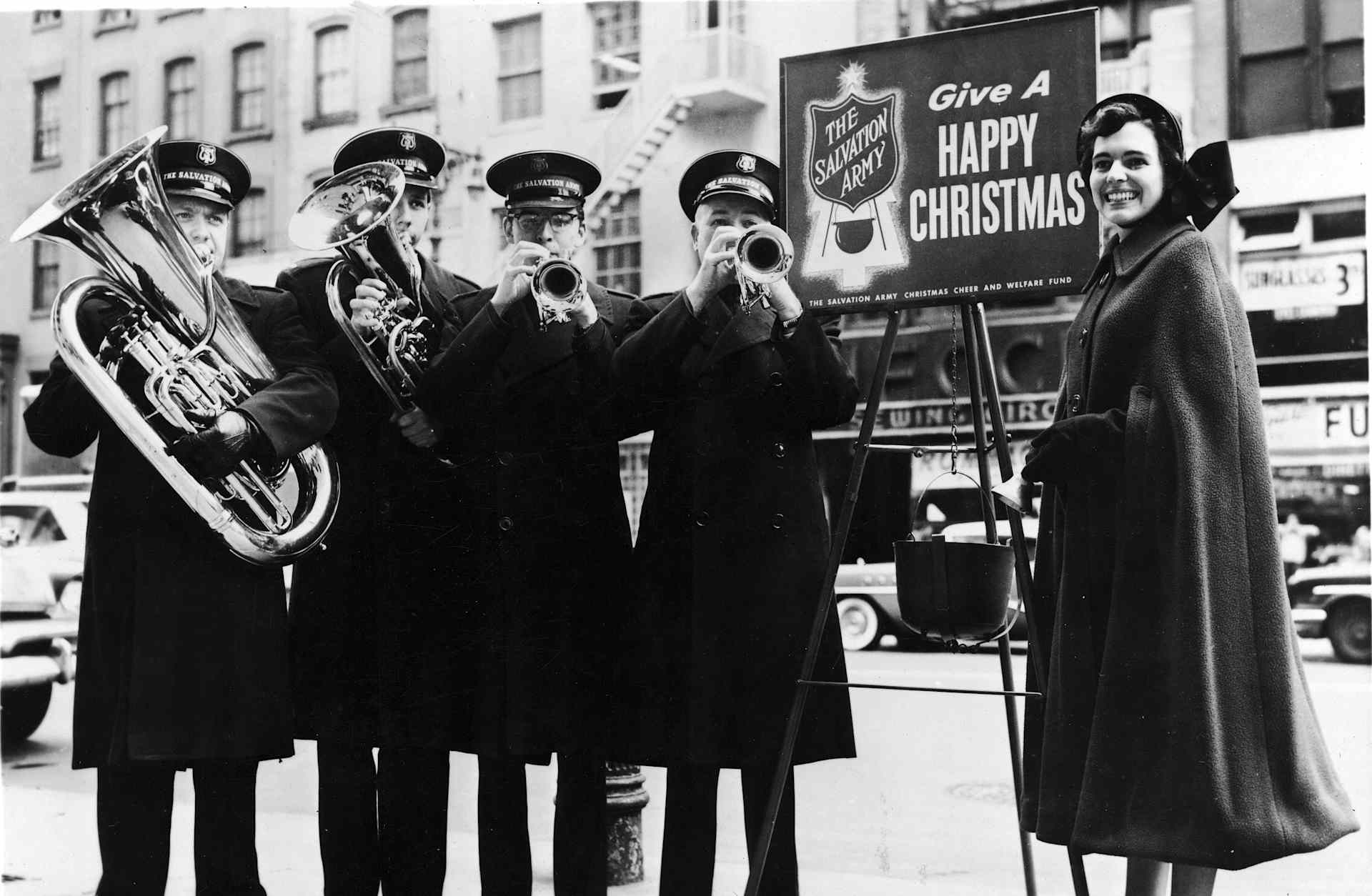 Four Salavation Army brass musicians play on a sidewalk while a female bell ringer stands by, New York City, circa 1950s.
