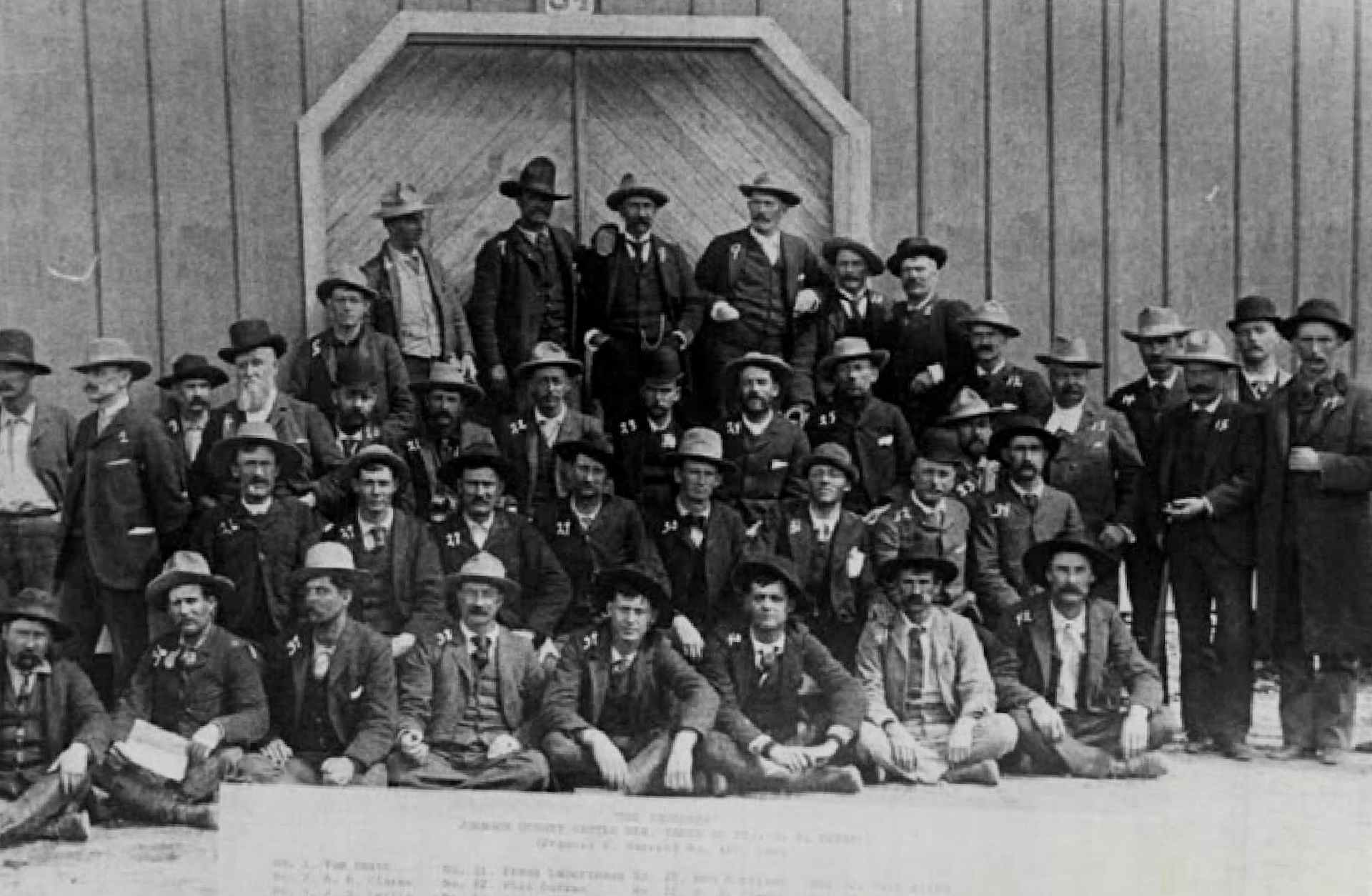 About 40 men in suits and hats posed in front of a barn. After this group of hitmen hired by Wyoming cattle barons to murder settlers were rescued from a siege by the cavalry, they were lined up for this photo. Due to influence from their wealthy backers, the gunmen were all eventually released.